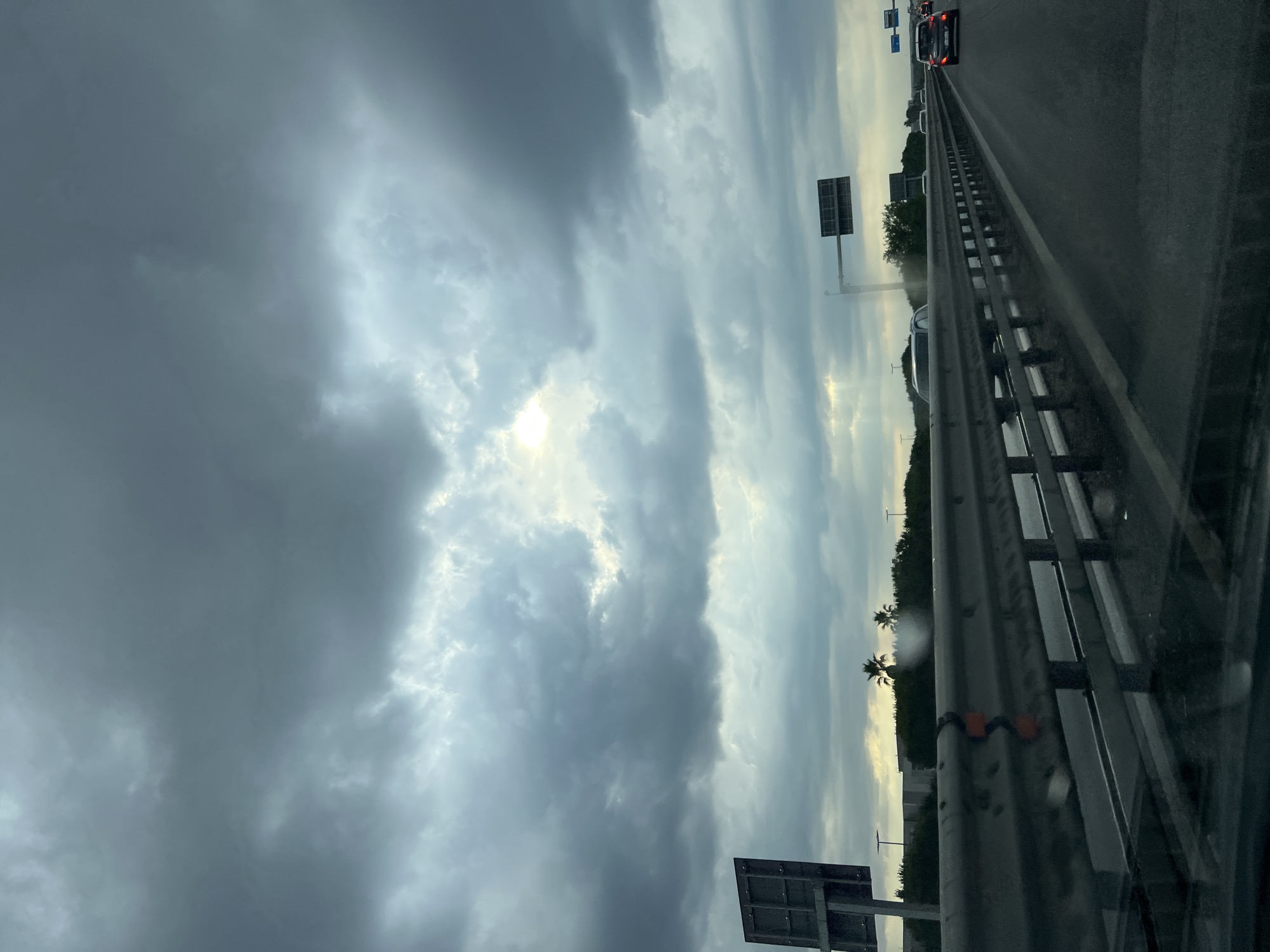 The image captures a view from a vehicle traveling on a highway in Molfetta, Italy, under a dramatic sky filled with heavy, dark clouds. Sunlight breaks through a gap in the clouds, casting rays of light onto the scene. The highway has a metal guardrail separating lanes of traffic. Palm trees and vegetation line the side of the road, with industrial or commercial buildings visible in the distance. Several vehicles, including cars and what appears to be a truck, are traveling on the highway. A large, dark rectangular sign is mounted on a pole on the left side of the road, and other traffic signs are visible further down the highway. The overall atmosphere suggests an impending storm.