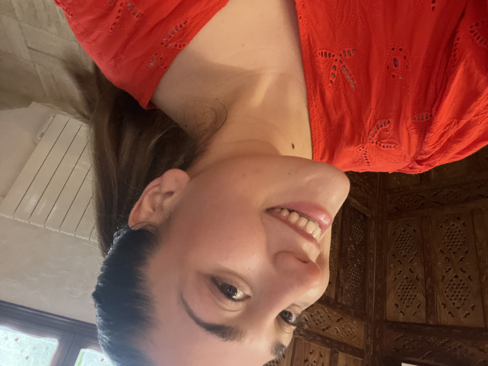 A young adult female is depicted in a close-up, selfie-style photograph taken indoors in Ancenis, France. She has fair skin, dark hair pulled back into a ponytail, and is seen smiling with teeth exposed, looking towards the camera. A small dark mole is present on the lower left side of her neck. She is wearing a bright red top featuring embroidered eyelet details on the sleeves and neckline. The background to her left consists of an intricately carved dark wooden room divider or screen. To her right, a light-colored wall is visible, featuring a white panel radiator and a window with multiple panes, through which blurred greenery can be observed. The scene is illuminated by indoor lighting.