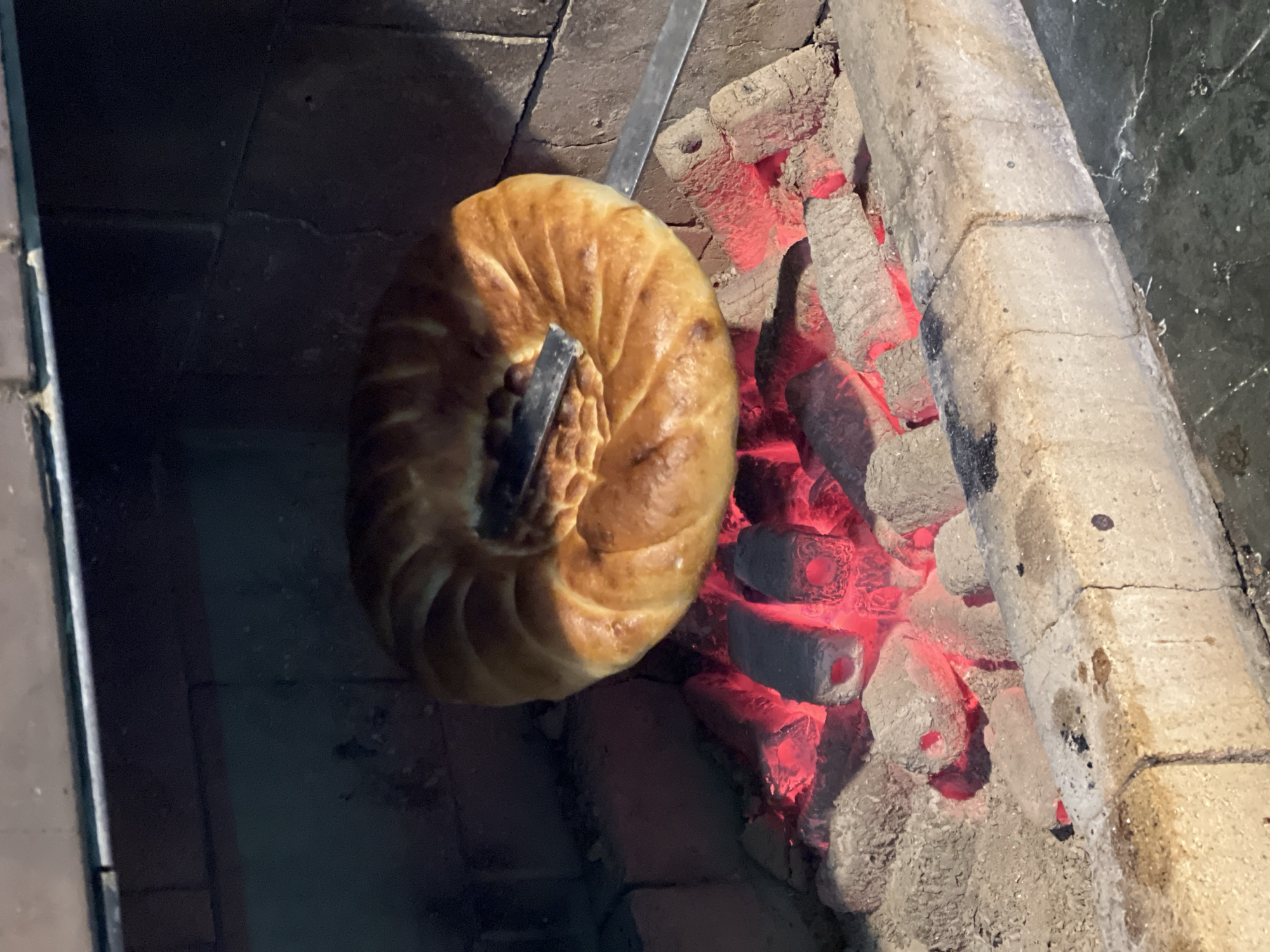 A round, golden-brown bread, possibly a type of flatbread or lavash, is being cooked over glowing red coals in a brick oven. The bread has a decorative, swirled pattern on its surface. A metal skewer or tool is inserted through the center of the bread, holding it in place over the heat. The oven interior is dark, with visible brickwork. The foreground shows the edge of the oven and the hot coals. The scene appears to be a traditional cooking method, possibly in a restaurant or home kitchen in Listvyanka, Russia, known for its culinary traditions and proximity to Lake Baikal. There are no people visible in the image, and the focus is entirely on the bread and the cooking process.