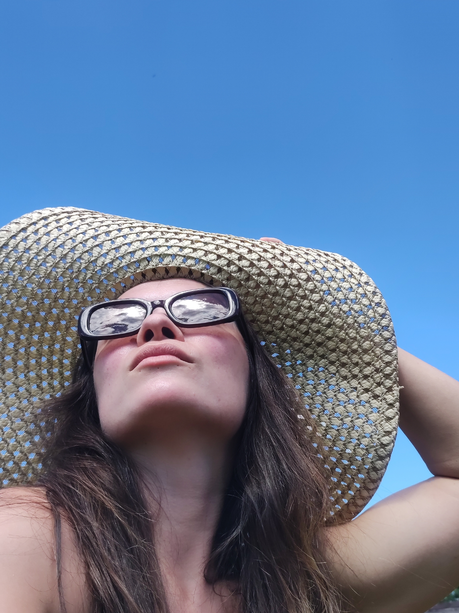A low-angle, close-up selfie of a woman wearing a straw hat and sunglasses, looking up at the sky. The woman has long brown hair and her face is partially obscured by the hat. The sunglasses reflect the sky and clouds. The background is a clear blue sky. The image appears to be taken outdoors on a sunny day. There is no discernible location in the city of Фоки, Russia, based on the visual information provided.