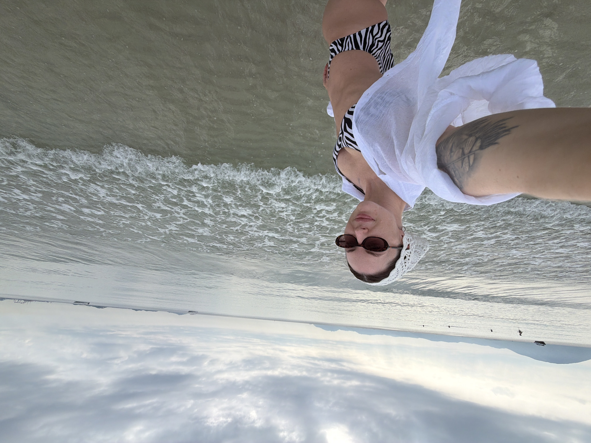 A woman with a white headscarf and sunglasses takes a selfie at the beach. She is wearing a black and white zebra-striped bikini and a white sheer shirt. The ocean is calm with small waves, and the sky is overcast. In the background, there are hills and a few boats on the water. The scene appears to be in Dong Ninh Hoa Ward, Vietnam.