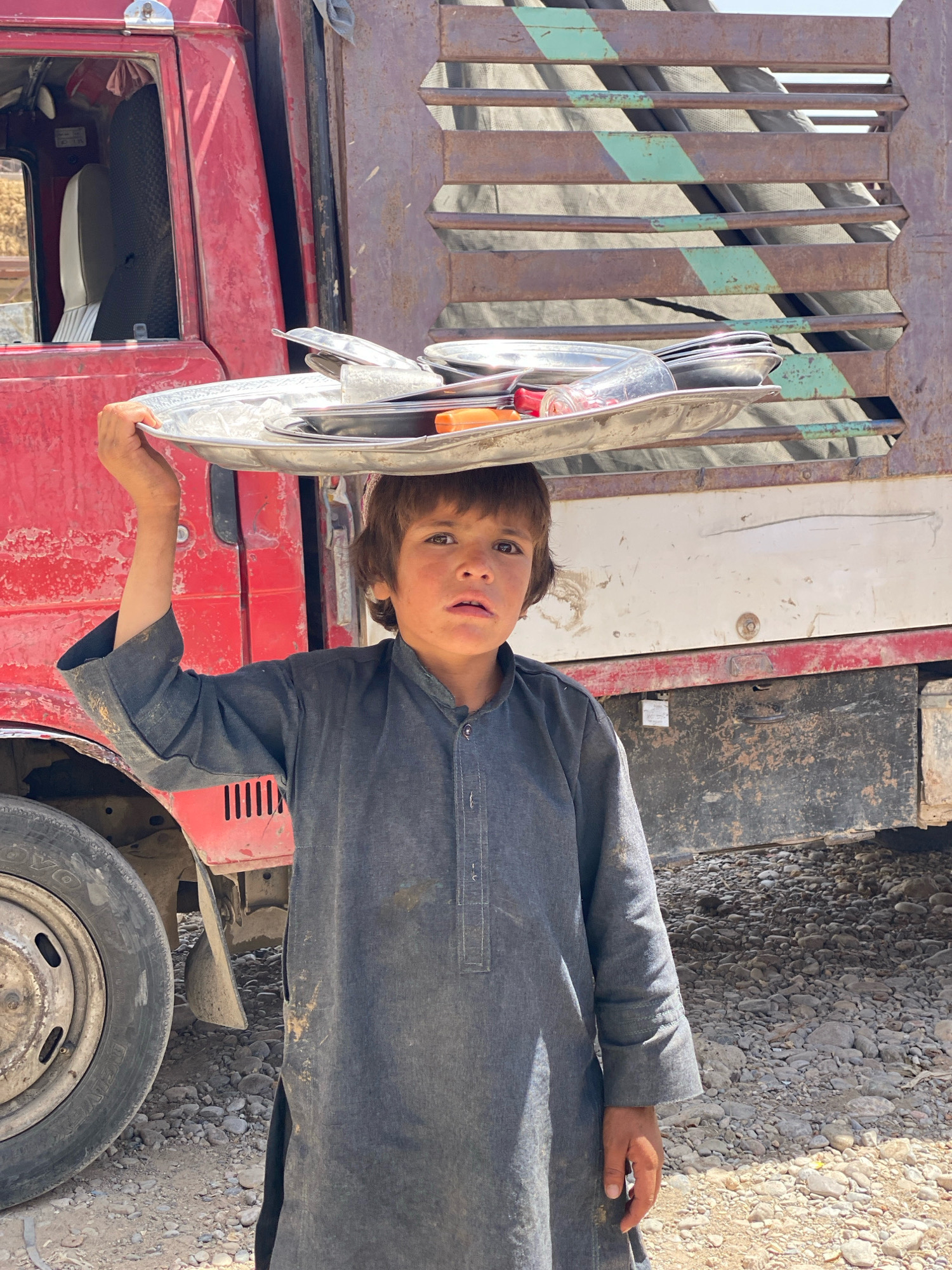 A young boy stands in front of a red truck in a dusty, rocky outdoor environment, likely in Kandahar, Afghanistan. The boy is carrying a large metal tray on his head, which is filled with various metal items, including plates, bowls, and a glass. He is wearing a dark grey traditional garment. The truck is large and appears to be a commercial vehicle, with a rusty metal cargo area. The ground is covered in small rocks and dirt. The overall scene suggests a rural or semi-urban setting with a focus on the boy and his task.