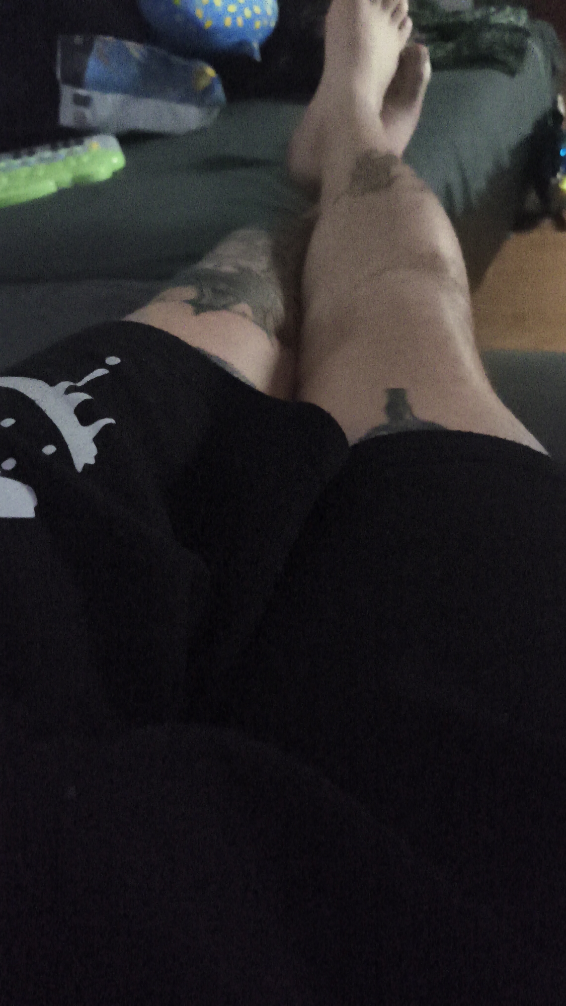 The image depicts a person's legs resting on a dark-colored couch. The person is wearing black shorts with a white design on the left leg. Both legs have tattoos. One leg is crossed over the other. The person's bare feet are visible, resting on the couch. The background includes other items on the couch such as toys or containers. The specific location in Nowy Tomyśl, Poland is not discernible from the image.