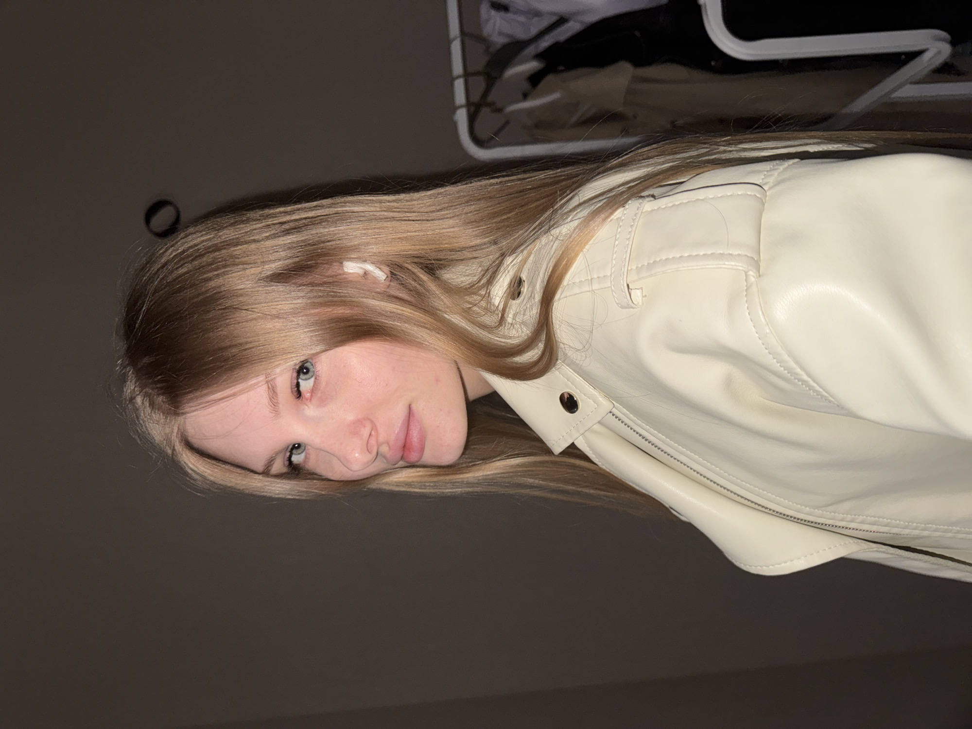 The image displays a head-and-shoulders portrait of a young woman, taken indoors in Perm, Russia. The photograph is oriented sideways, with the subject's head positioned on the left side of the frame. The woman has long, straight blonde hair that flows around her face and over her shoulders. Her blue eyes are directed towards the camera, and she has a calm, neutral expression with a slight hint of a smile.
She is wearing a light cream or off-white leather-look jacket, featuring visible stitching, a silver-toned zipper, and metallic snap buttons on the lapel. A small white earbud is visible in her left ear. The background behind her is largely dark and indistinct, suggesting low light conditions. In the upper right corner of the frame, a metal clothes rack with some dark garments hanging on it is partially visible, indicating an indoor setting, possibly a room or closet. A dark, circular object, which appears to be a hair tie, is faintly visible against the dark wall above her head. No text is visible in the image.