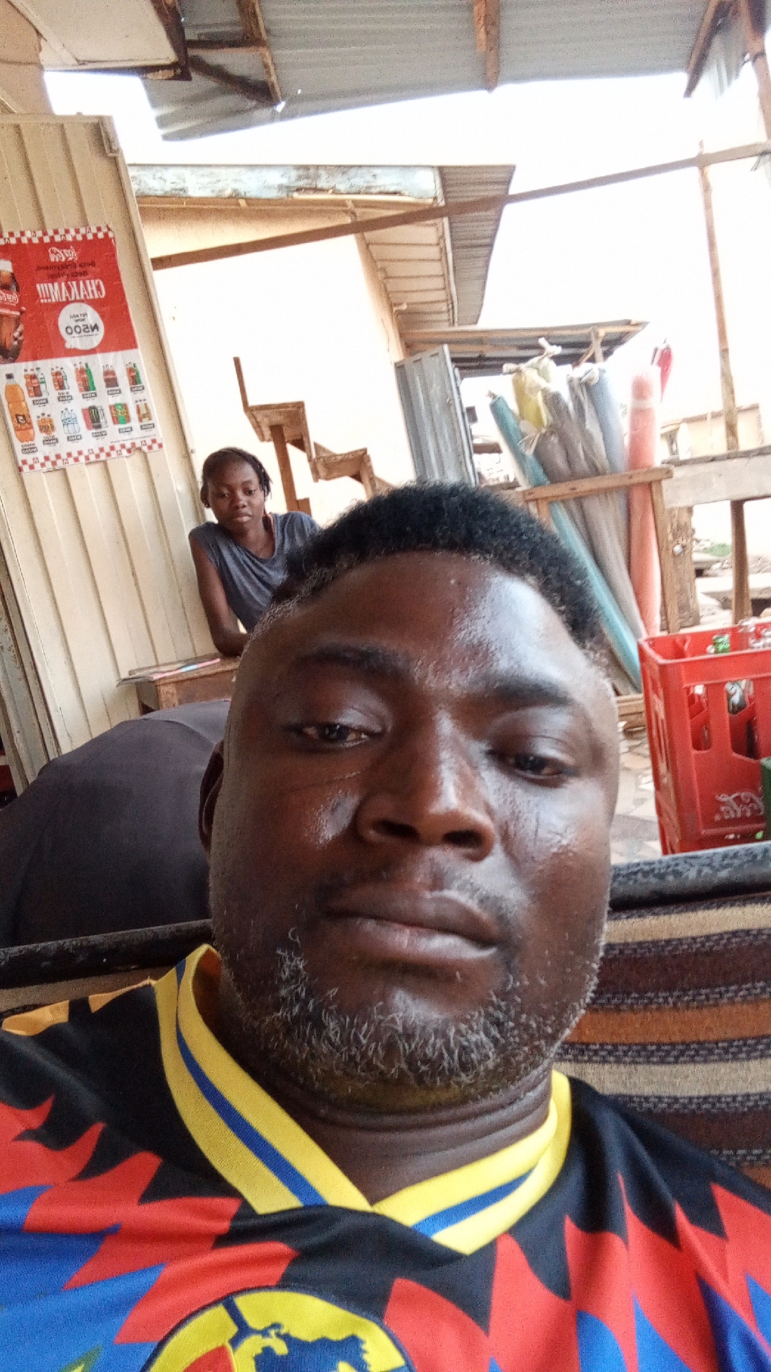 The image features a selfie-style portrait of a man in the foreground, with a woman and an informal shop setting visible behind him in Sabon Tasha, Nigeria.
The man in the foreground has dark skin, a short dark haircut, and a graying beard and mustache. He is wearing a distinctive black shirt with bold red, yellow, and blue geometric patterns, possibly a sports jersey. His expression is neutral as he looks directly into the camera.
In the mid-ground, a young woman with dark skin and dark hair is partially visible, leaning on a surface. She wears a plain grey t-shirt and looks towards the camera.
The background depicts a rustic commercial environment. On the left, a white poster, partially upside down and mirrored, advertises beverages with the prominent text "CHAKAAA!!!" and "N500," likely indicating a price in Nigerian Naira, alongside illustrations of various bottled drinks. The setting includes wooden beams, corrugated metal sheets forming a roof structure, and a light-colored building wall. On the right, a red plastic crate, typically used for bottles, is visible next to a collection of rolled-up items, possibly mats or fabrics, leaning against a wooden support.
The scene is illuminated by bright daylight, suggesting it is daytime. The overall ambiance is that of a casual, local shop or stall.