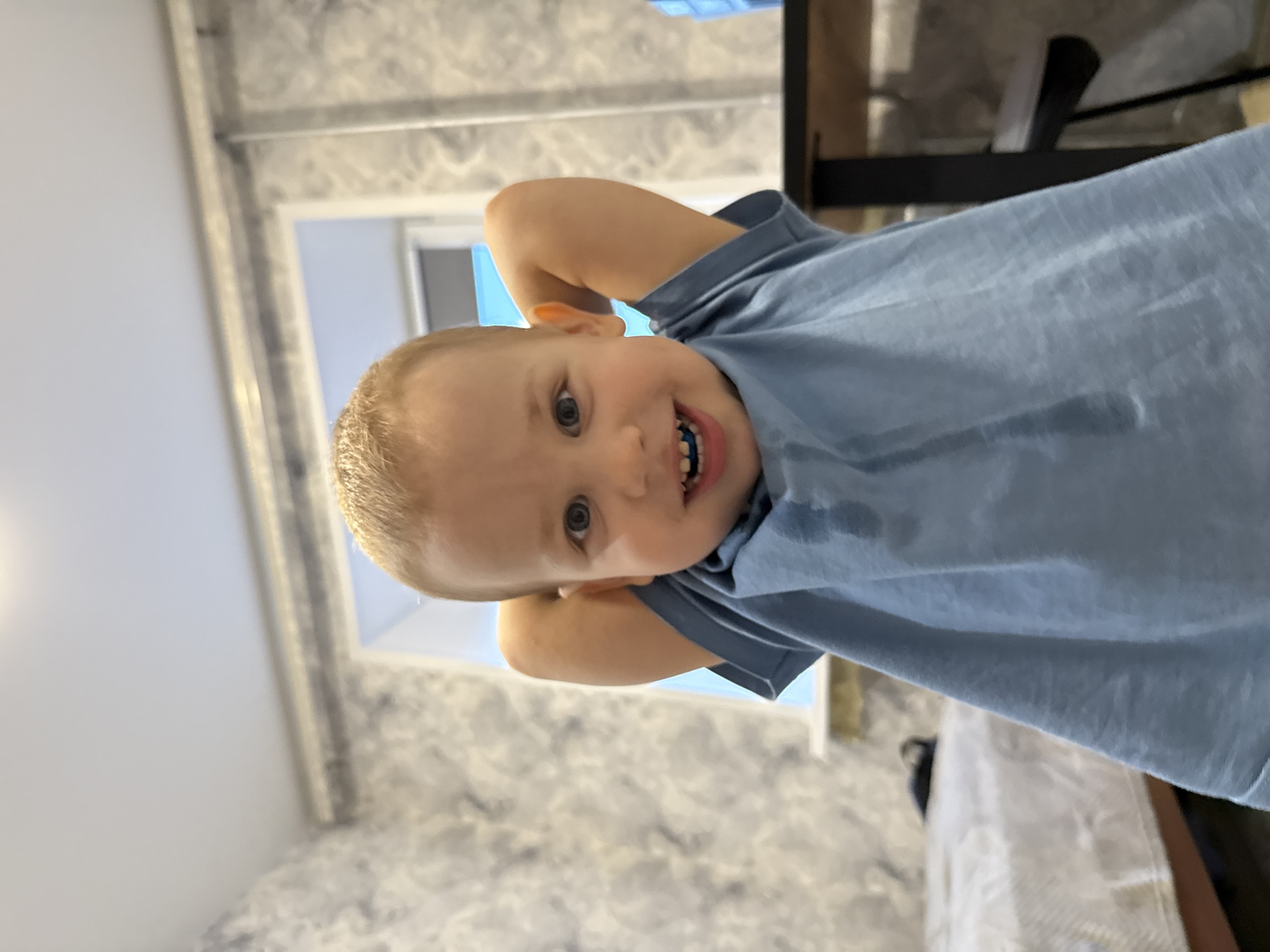 A young boy with blond hair and blue eyes is smiling at the camera, with his hands behind his head. He is wearing a blue t-shirt. The background shows a room with patterned wallpaper, a window, and part of a desk with a chair. The location is Rusanovo, Russia.