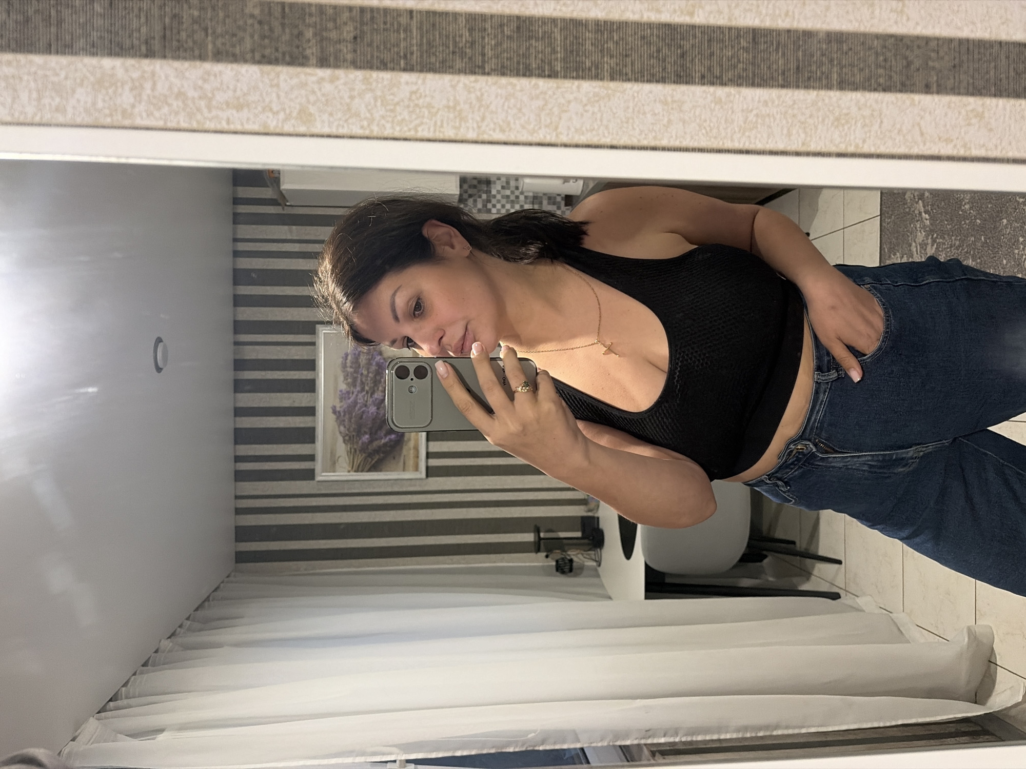 The media file is a mirror selfie taken by a woman indoors. She is wearing a black tank top and blue jeans, with a gold necklace. She is holding a smartphone in her right hand and has her left hand in her pocket. In the background, a framed picture of lavender is visible on a wall with striped wallpaper. A white curtain hangs on the left side of the frame. The lighting suggests it is daytime. The setting appears to be a room, possibly a bathroom or bedroom. The woman has a slight smile on her face, suggesting a casual and relaxed mood. There is no visible text in the image. The location context is Sochi, Russia.