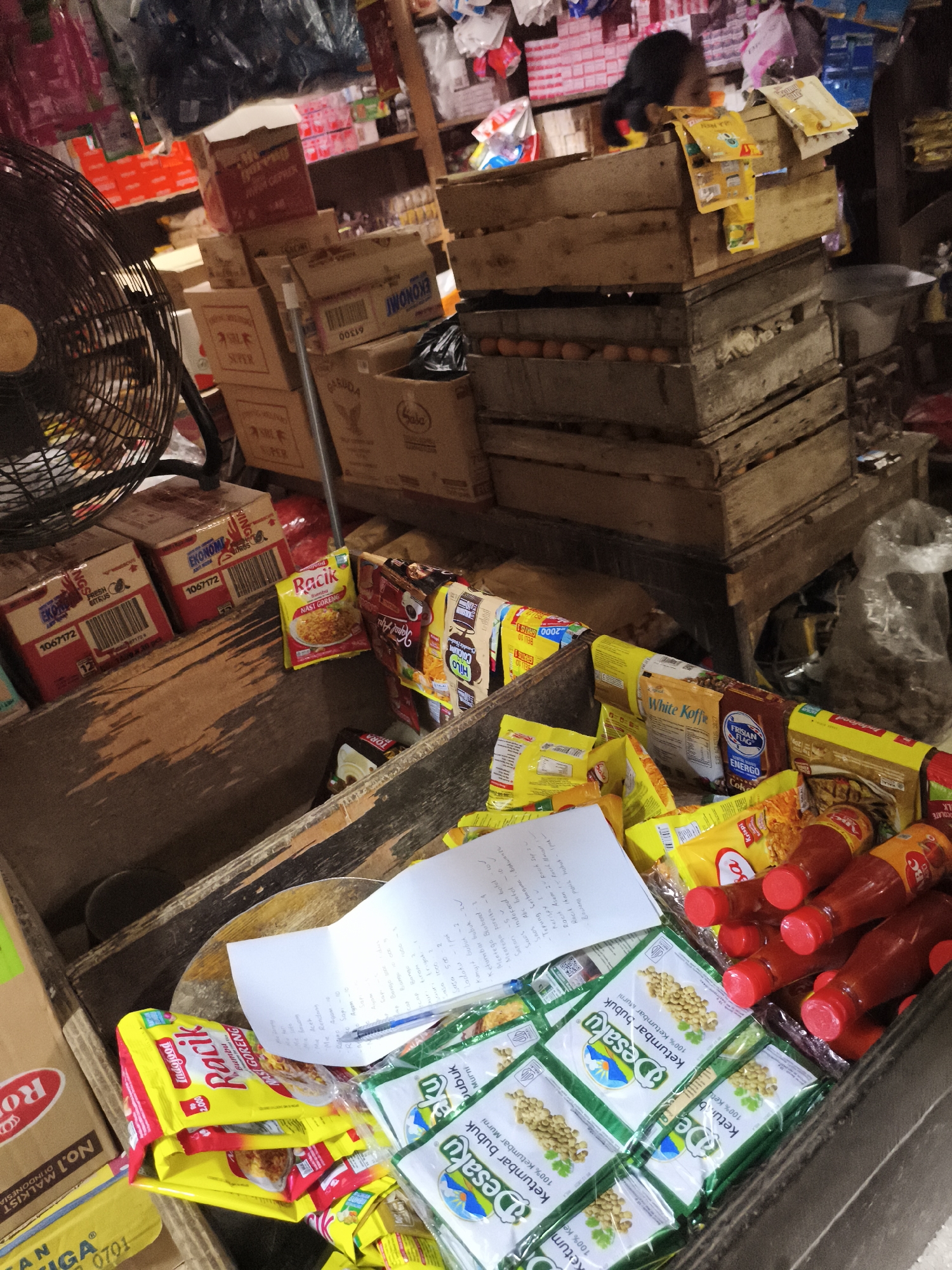 The image captures a cluttered scene inside a small shop, likely in Depok, Indonesia, given the brands and product labels visible. The focus is on a wooden crate filled with various food products, including packets of "Desaku Ketumbar Bubuk" (coriander powder), "Racik" seasoning, bottles of "ABC" chili sauce, instant coffee, and boxes of "Ekonomi" milk. A piece of paper with handwritten notes and a pen rest on top of the goods, suggesting an inventory list or sales record. In the background, stacks of wooden crates and cardboard boxes create a sense of cramped storage space. An electric fan stands to the left, and glimpses of other merchandise and a person are visible in the background, indicating a bustling retail environment.