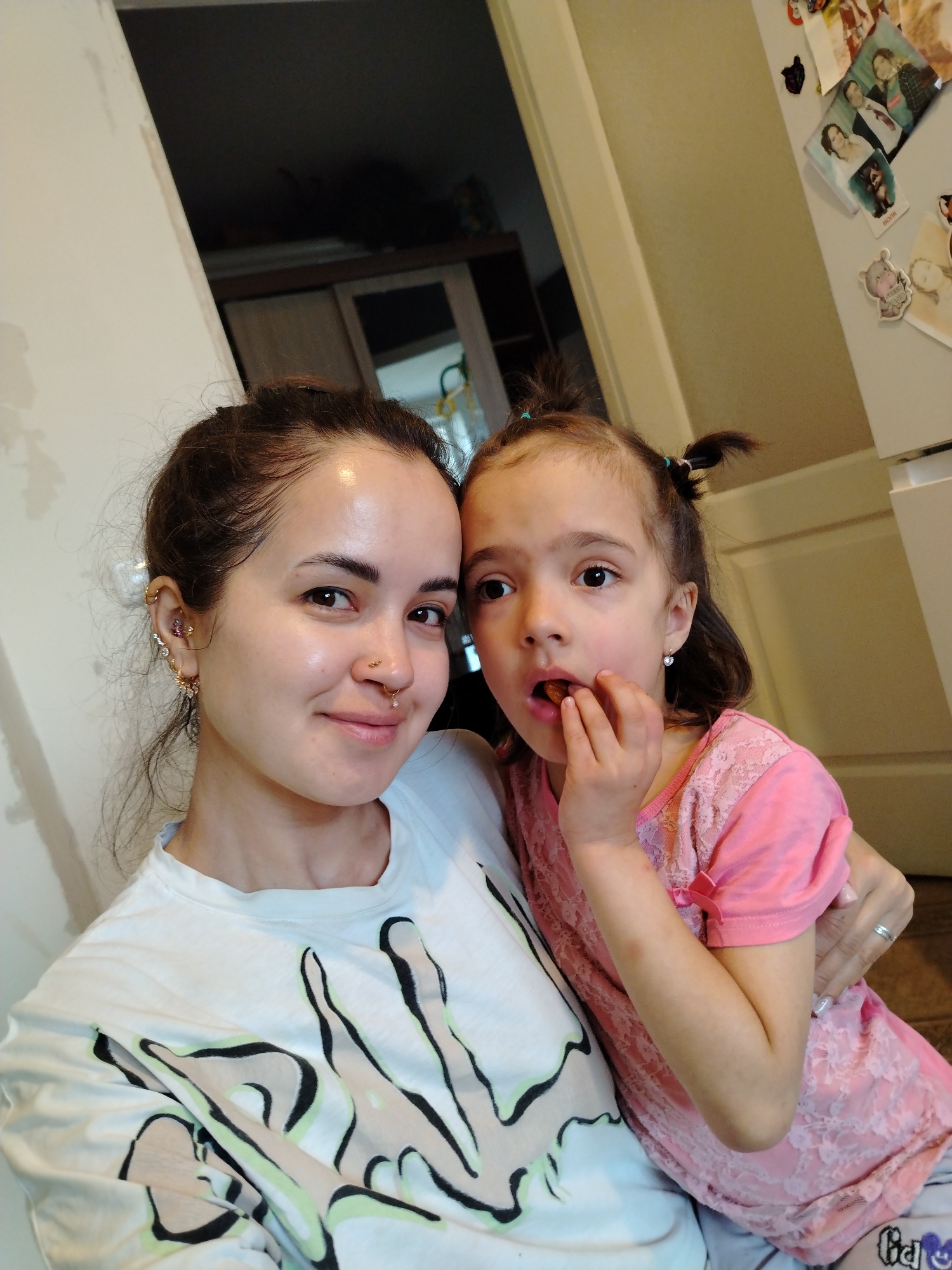 This indoor portrait features an adult woman and a young girl, likely a mother and daughter, in a close-up view within a home in Noviy, Russia. The background includes a light-colored wall with some texture on the left, and a refrigerator adorned with various magnets and photos (one clearly visible with the word "BOOK") on the right. A doorway or opening is also visible in the upper background.
The woman, positioned on the left, has dark hair tied back and multiple piercings, including ear piercings (hoops, helix, conch, daith) and facial piercings (septum and nostril rings). She wears a white t-shirt with a distinctive graphic design in light green, black, and beige. She looks directly at the camera with a soft, gentle smile.
To her right, the young girl has dark hair styled in two small ponytails. She is dressed in a pink top, which appears to have lace detailing. She is captured in the act of eating, holding a small piece of food to her mouth with her fingers, and her gaze is directed slightly away from the camera. She wears small silver stud earrings. A silver ring is visible on the adult woman's left hand, which is placed behind the girl. The scene conveys a candid and intimate moment between the two.