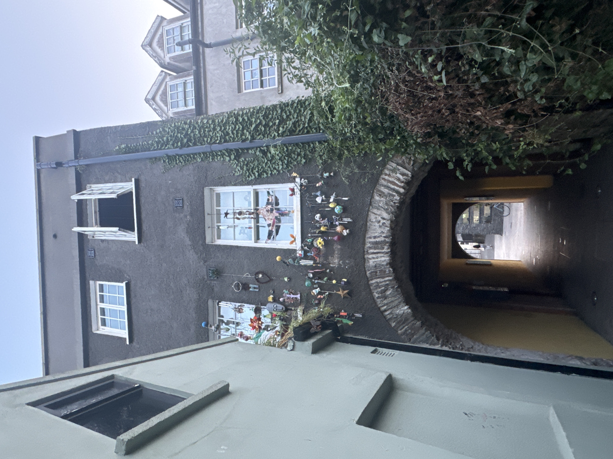 This is a low-angle, exterior shot of an archway leading into a narrow street in Waterford, Ireland. The archway is constructed of stone and is flanked by buildings. The building to the left is light green and has a slanted window. The building to the right is dark gray and has several windows, some of which are open. The facade of the dark gray building is adorned with numerous decorative items, including masks, chimes, and other hanging ornaments. Ivy covers a portion of this building and the adjacent building to the right, which is lighter in color and features dormer windows. Beyond the archway, the street is visible, with a person walking away from the camera and a bicycle parked. The sky is clear and pale blue.