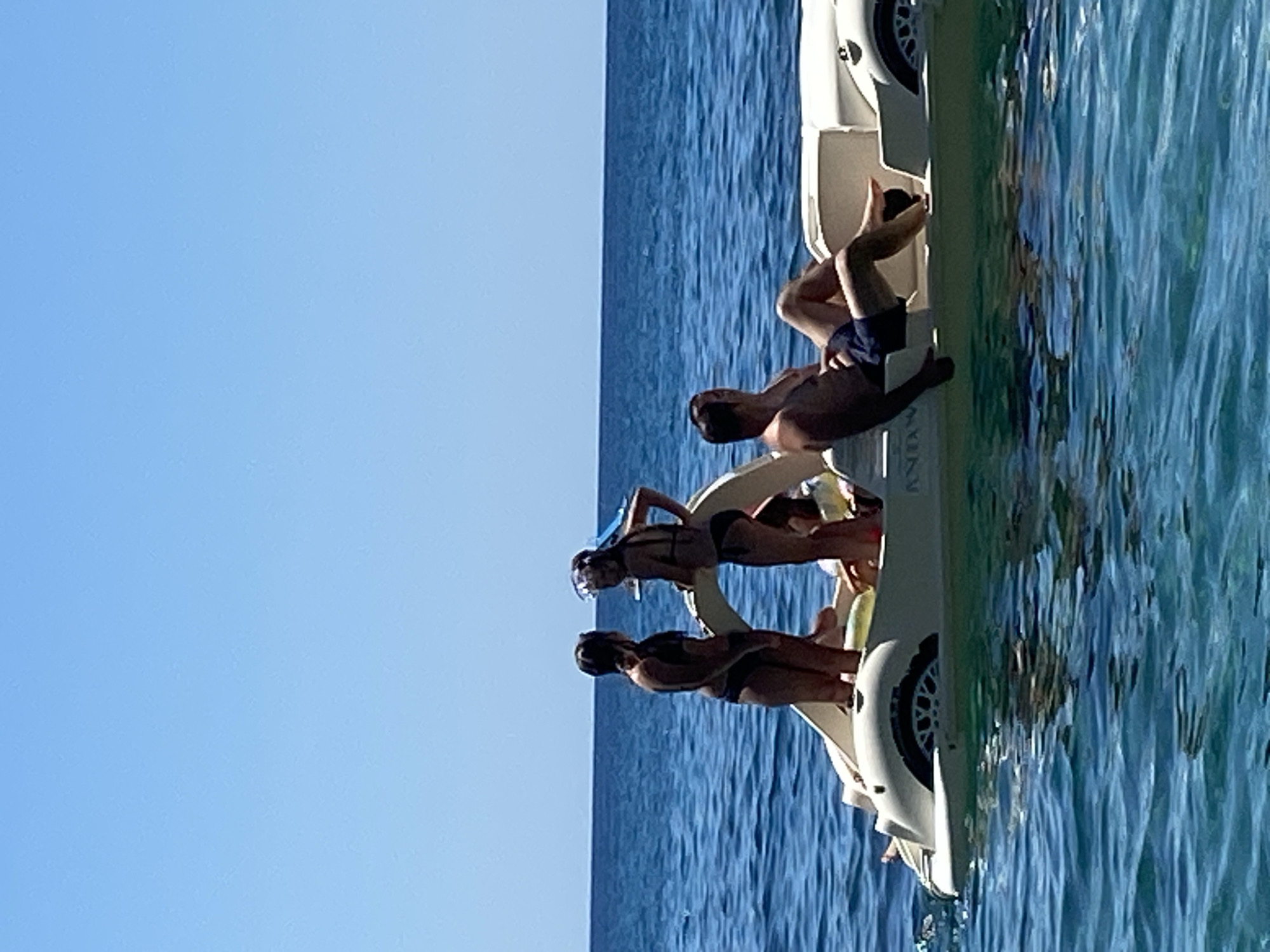 In the city of Prijevor, Montenegro, on a bright, clear day, a group of four individuals enjoys a leisurely ride on a white pedal boat. The boat, shaped like a compact convertible, floats on the calm, blue waters of the Adriatic Sea. Two women, wearing bikinis, stand near the front of the boat. One woman, facing away from the camera, has her hair pulled back. The other woman, facing the camera, wears a snorkeling mask pushed up on her forehead. In the rear of the boat, a man is seated, facing left and looking upwards. Next to him, another person, partially obscured, is also seated on the boat. The boat has a visible wheel on its side and the word "ANNA" is faintly visible on its hull. The horizon line between the sea and the sky is clearly defined.