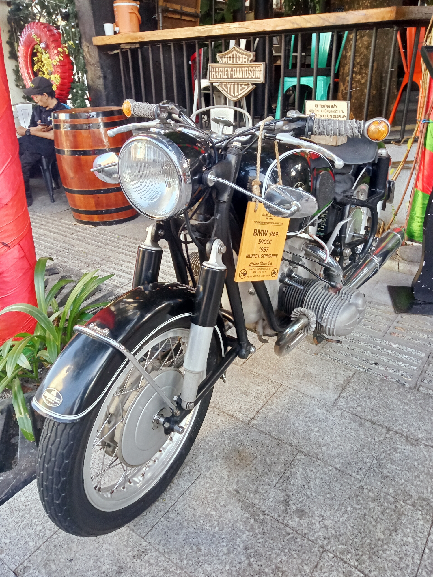 The image depicts a vintage BMW R69 motorcycle, 590cc, model 1957, manufactured in Munich, Germany, displayed at Palms Nha Trang, Vietnam. A sign attached to the motorcycle reads "THE VINTAGE MOTORCYCLE COLLECTION," "Classic Never Die," and has a warning in Vietnamese and English ("PLEASE DO NOT SIT ON OR TOUCH"). In the background, a wooden barrel is visible. A man is seated, looking at his phone. Also visible in the background are a Harley-Davidson sign, plants, and colorful decorations.