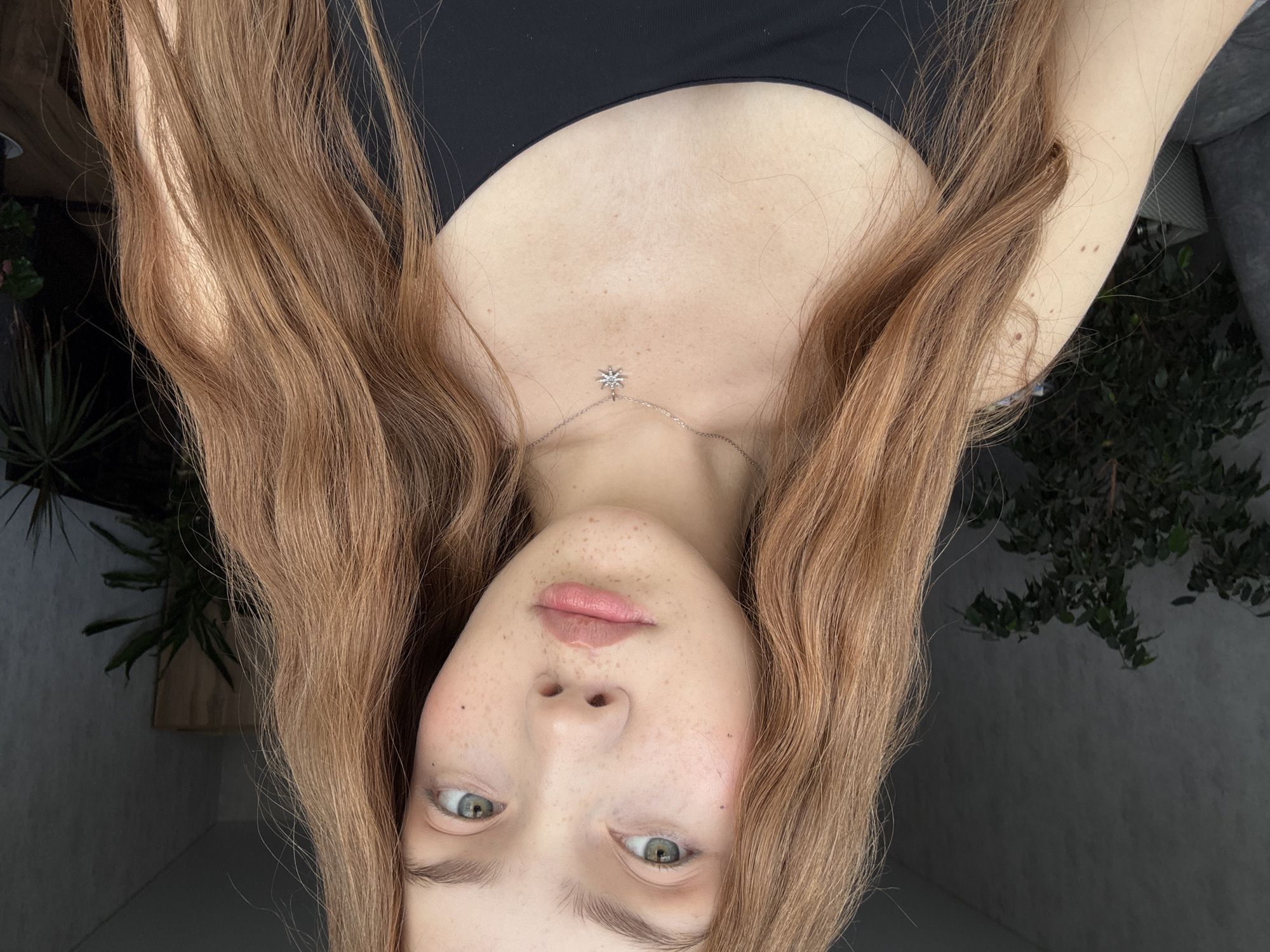 An upside-down close-up portrait features a person, likely a young adult female, with long, wavy, reddish-brown hair. Her face is positioned towards the bottom of the frame, with her light-colored, greenish-blue eyes looking upwards. Prominent freckles dot her nose and cheeks, and her lips are naturally pink and slightly parted. She is wearing a dark, possibly black, low-cut top that reveals her upper chest. A delicate silver-toned necklace featuring a small, star-shaped pendant is visible on her décolletage. Her long hair cascades upwards, framing her face and body.
In the background, visible on both sides of her hair, are glimpses of dark green foliage from plants, suggesting an indoor setting with large windows or an outdoor area such as a balcony. The lighting is natural and bright, indicating daytime. The person’s expression appears neutral and calm. There is no visible text in the image.