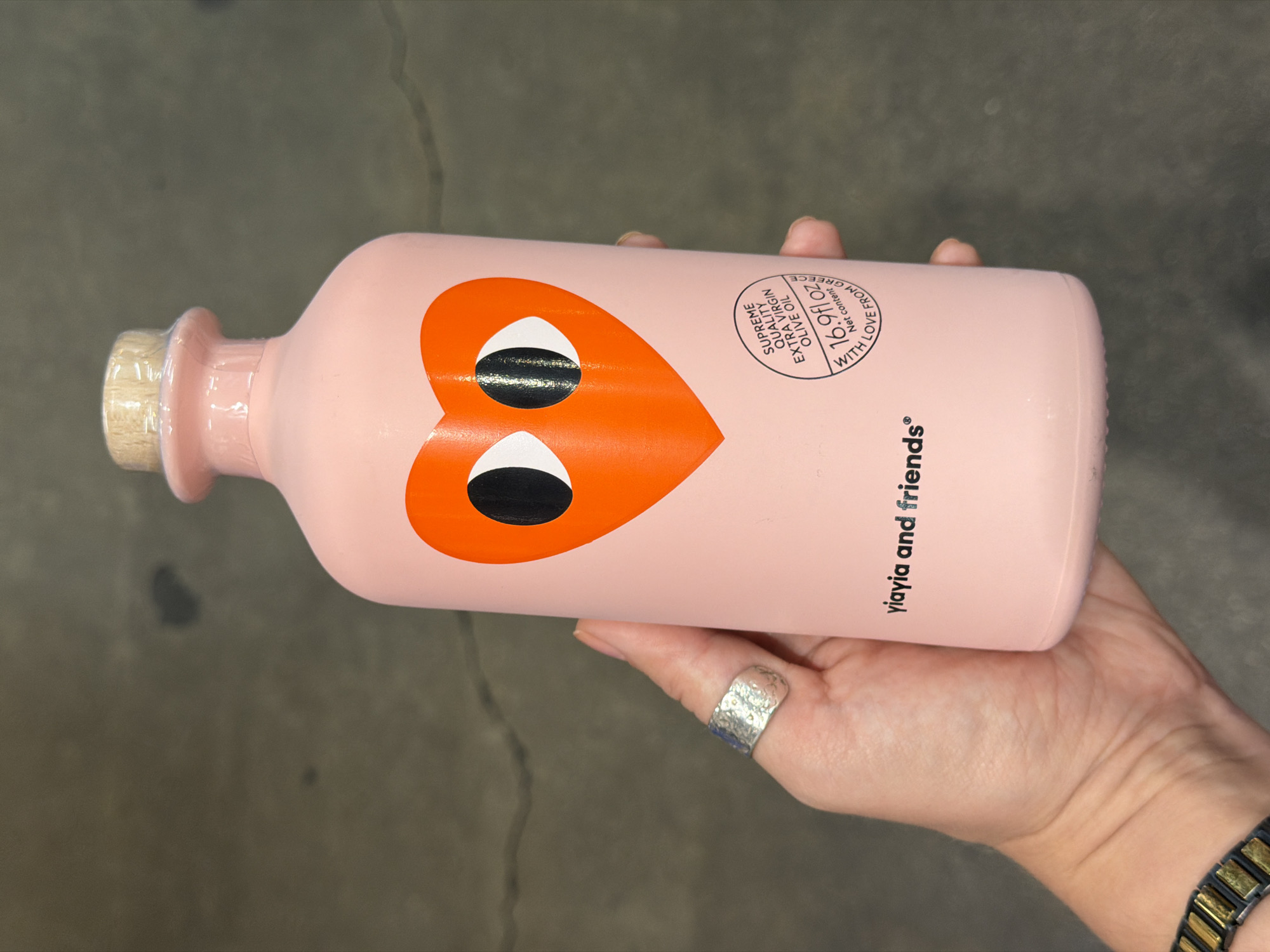 Here is a description of the image:
A person's hand is holding a pink bottle of YiaYia and Friends brand olive oil. The bottle features a stylized orange heart design with large black eyes. The label indicates the product is "Supreme Quality Extra Virgin Olive Oil, 16.9 fl oz, with love from Greece." The bottle has a light brown wooden-like cork at the top. The person's hand is wearing a silver ring on their thumb and has a watch visible at the wrist. The background is a gray surface.