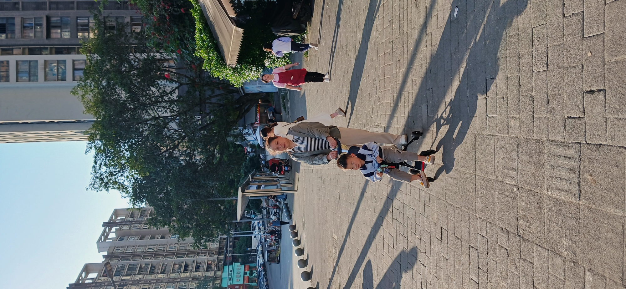 The image depicts a paved public space in Jiulongpo District, China. An adult woman, dressed in casual clothing, is pushing a young boy on a tricycle. Other people are visible in the background, including a woman in a red apron. The area is urban, with buildings and trees lining the walkway. The ground is tiled and shadows from the figures are visible. A street with parked cars and scooters can be seen, along with round bollards.
