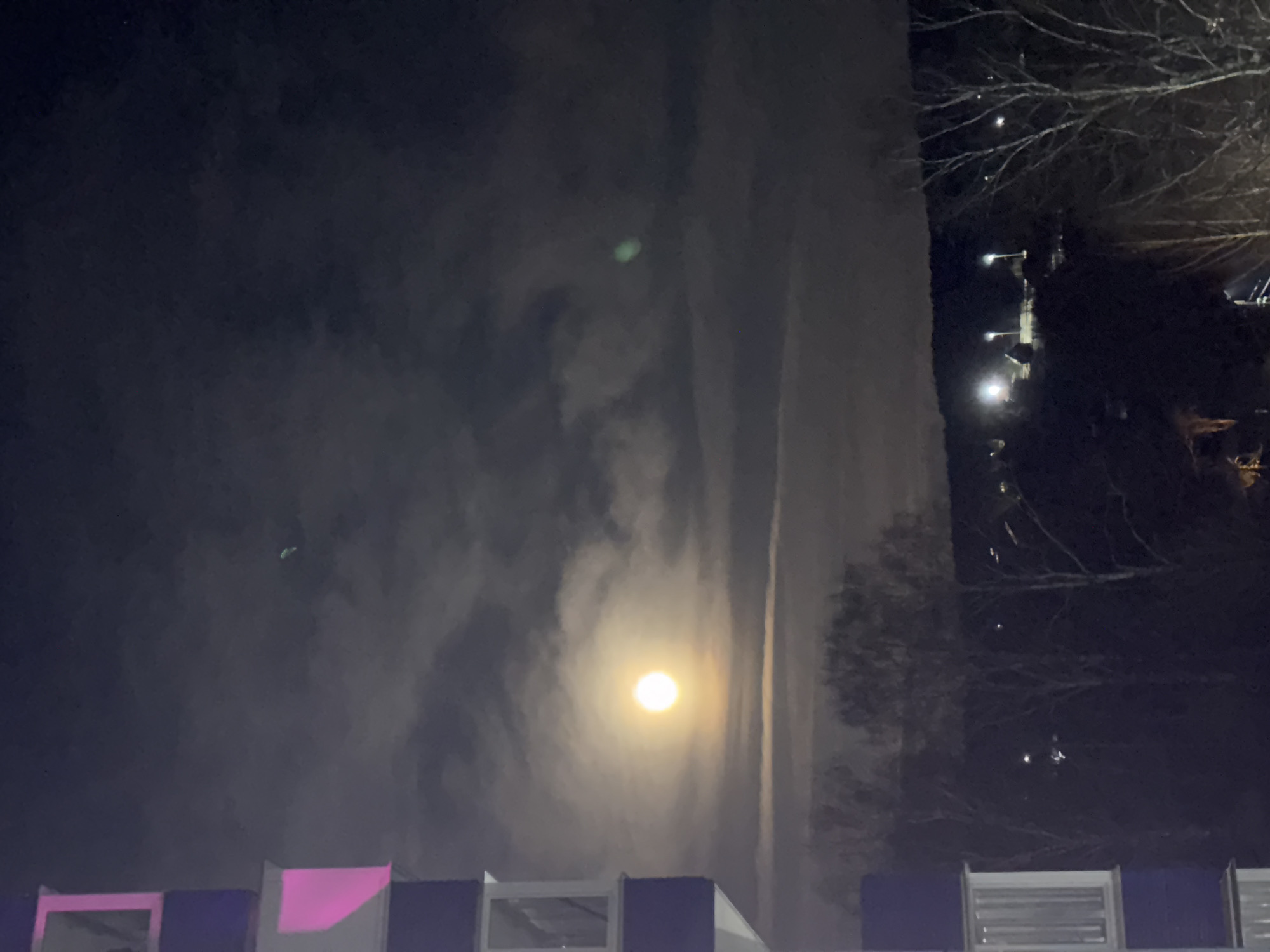 The image, taken at night, features a bright moon partially obscured by clouds. Trees are visible in silhouette on the right side of the frame, along with distant streetlights. The foreground shows a building structure with elements colored pink, white, and dark blue. The location is identified as Chaikovsky, Russia. The photo is taken from a low vantage point, looking upwards.