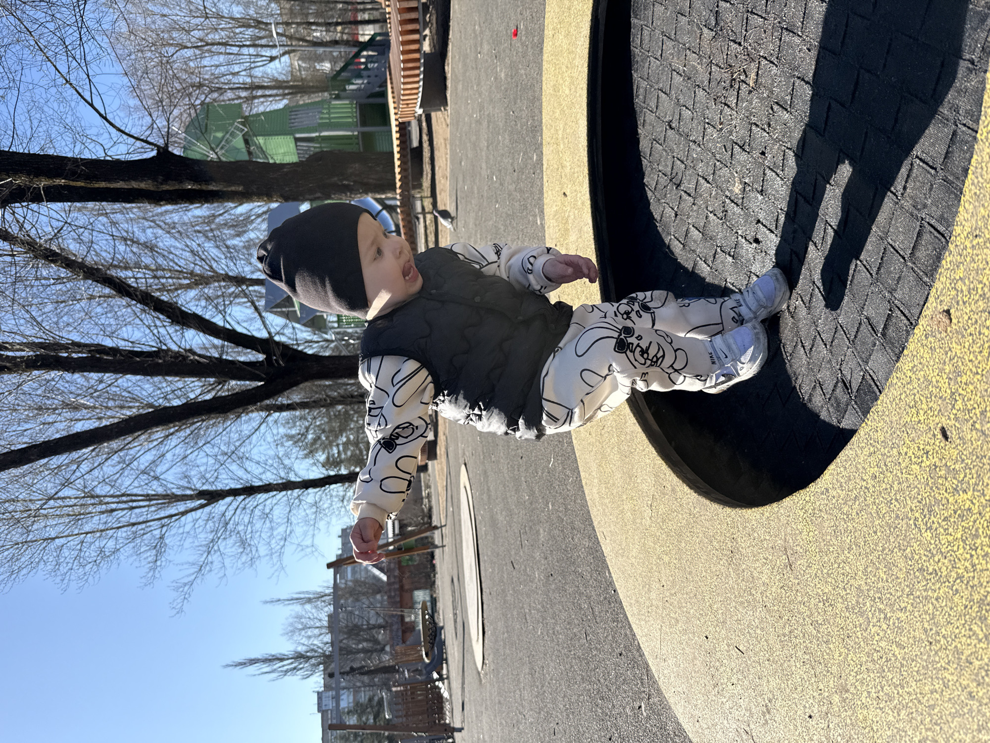 A rotated eye-level outdoor shot depicts a young child in a black beanie, white patterned sweat suit and black vest seated on playground equipment. The child faces left with their mouth open. In the background is a park with trees. The sky is blue.