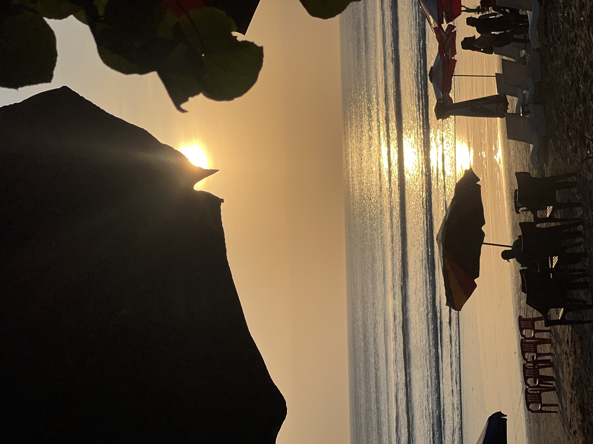The image depicts a serene beach scene in Kuta, Indonesia, during sunset. The sun, a golden orb, is partially obscured by dark foliage in the foreground, casting a warm, ethereal glow across the sky and the ocean. Gentle waves roll onto the sandy shore, reflecting the sun's shimmering light. Beach chairs and umbrellas are scattered across the sand, some occupied by silhouetted figures enjoying the tranquil evening. The overall atmosphere is peaceful and idyllic, capturing the essence of a tropical sunset.