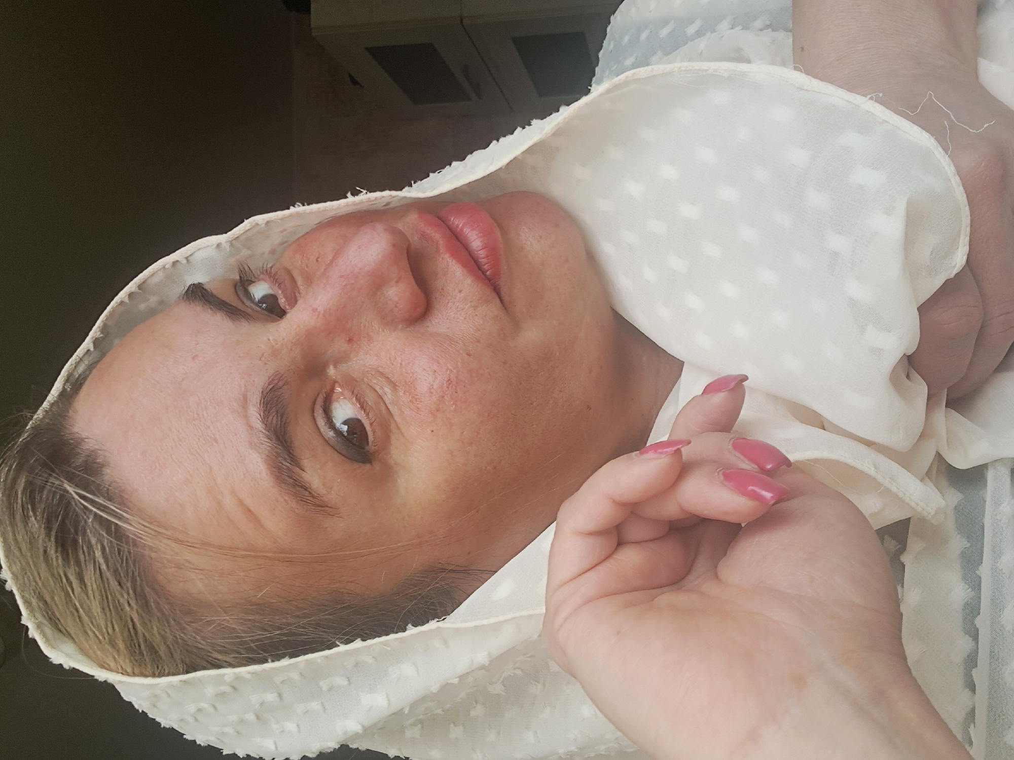 A close-up, rotated portrait shows a woman indoors, possibly in Elektrostal, Russia. She is lying down, looking directly at the camera, with her head tilted to her right. Her face, hands, and shoulders are visible. She has light-colored hair, some of which peeks out from under a light cream or off-white head covering or shawl, which features a subtle, textured pattern of small, raised dots. Her lips are made up in a pinkish-red shade, and her eyes are open, with visible eyebrows. Her skin has some natural redness. One hand is near her face, holding a part of the fabric, and the other hand is seen further down. Her fingernails are painted with a bright pink polish. In the background, out of focus, are faint hints of indoor furnishings, including what appears to be part of a cabinet or shelf with dark openings. The overall lighting suggests an indoor setting during the daytime. There is no visible text in the image.