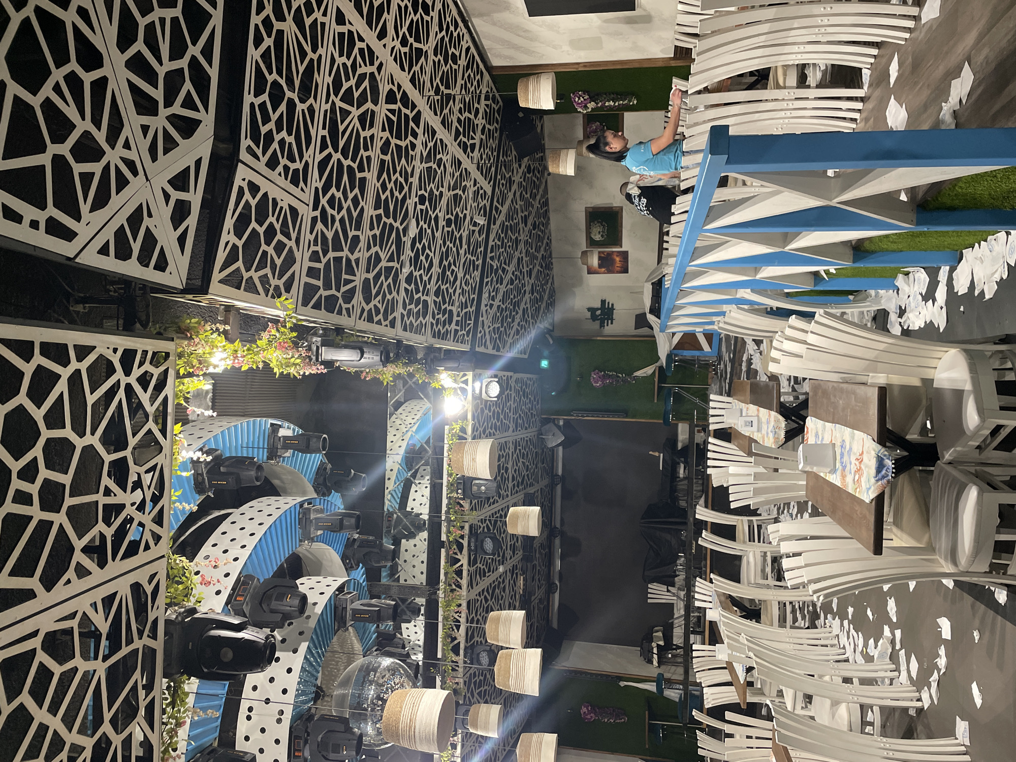 The image depicts an outdoor seating area, possibly a restaurant or event space, in Plovdiv, Bulgaria. The space is characterized by tables and white chairs. A partially visible blue railing with a geometric design is present. One female and one male person are visible, standing next to the railing. The background includes decorative walls with geometric patterns, spherical decorative elements, planters, lights, and greenery. The area shows traces of paper debris on the ground.