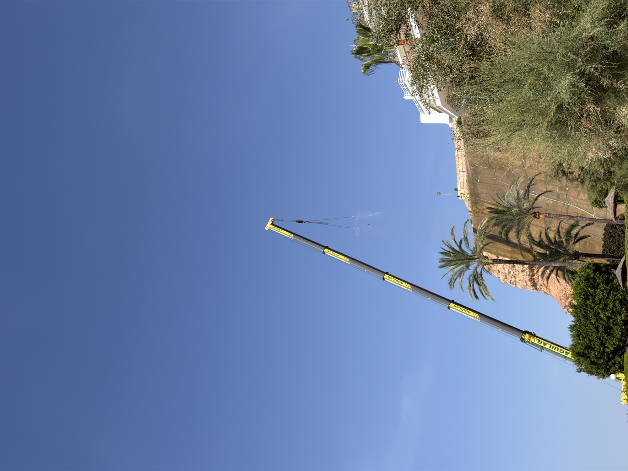 An outdoor daytime scene in Portals Nous, Spain, shows a large yellow and black crane extending diagonally from the lower left of the frame upwards into a clear blue sky. The crane is lifting a transparent, rectangular object. To the right of the crane, a steep, textured cliff face rises, partially covered with vegetation. At the top of the cliff, a white building with balconies is visible, surrounded by lush greenery and palm trees. The scene suggests construction or maintenance work taking place on the cliffside.