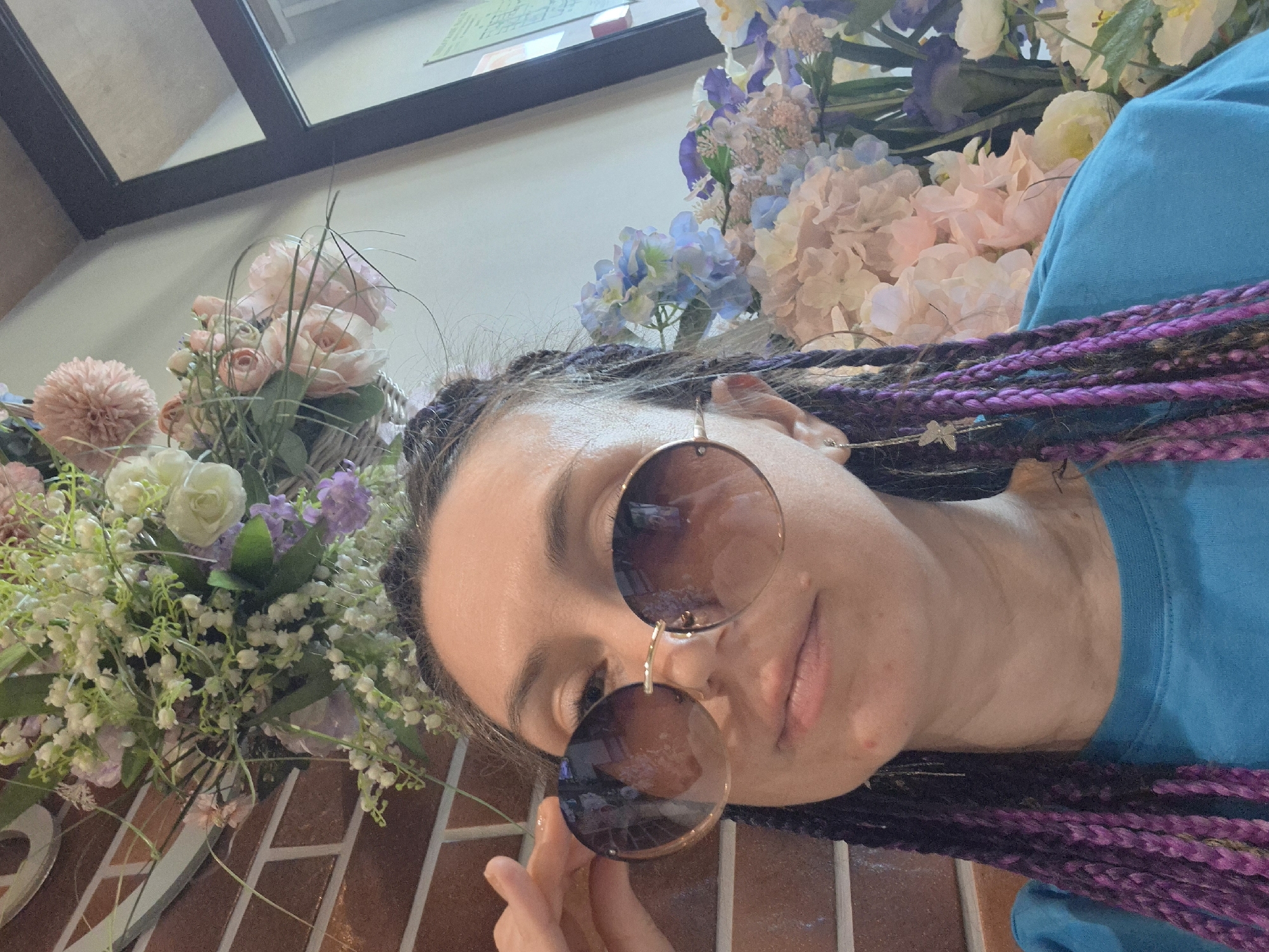 A woman with purple braids poses with round sunglasses in front of bouquets of flowers in Votkinsk, Russia. The lower part of a framed structure, possibly a window, is visible in the upper left. A brick-like surface is beneath the flowers on the left. The woman is wearing a bright blue shirt.