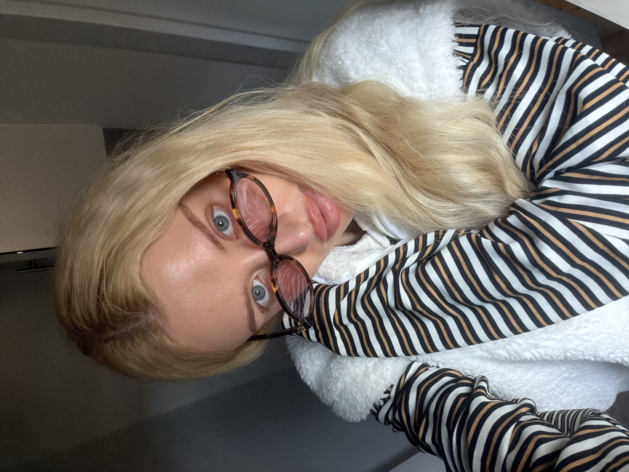 A woman with blond hair and glasses is depicted in the image. She is wearing a striped shirt with black, white, and tan stripes and a white, fuzzy jacket. The environment appears to be indoors, though specific details about the location in Belgrade, Serbia, are not evident.
