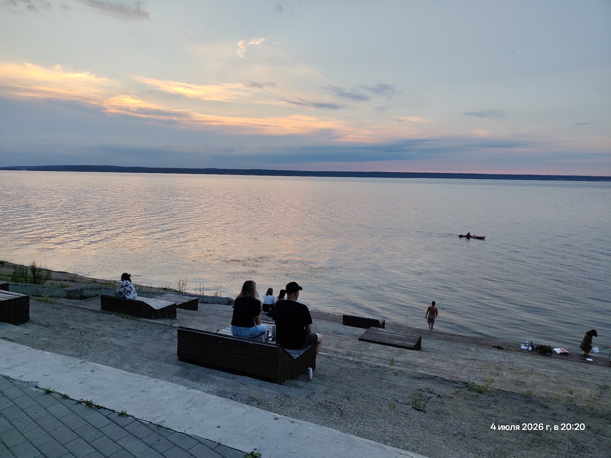 The image captures a serene evening scene at a waterfront in an unknown city. The sky is painted with the soft hues of a sunset, transitioning from warm oranges and yellows near the horizon to cooler blues and grays overhead, with wispy clouds scattered across the expanse. A vast body of water, likely a river or lake, stretches across the frame, its surface rippled and reflecting the sky's colors.
Along the embankment, several wooden benches are positioned, some occupied by people. In the foreground, a couple sits on a bench, facing the water, appearing to relax. To their left, another person is seated on a bench, also facing the water, with their back to the camera. Further to the left, a single individual is seated on a bench, further away.
On the water, a small boat with an occupant is visible in the distance. Closer to the shore, a person stands near the water's edge, holding a fishing rod. Several items, including bags and what appear to be bottles, are placed on the embankment near this individual. Another person is visible further down the embankment, near the right edge of the frame, standing by the water. The embankment itself is a mix of paved areas, gravel, and some sparse vegetation. The timestamp "4 июля 2026 г. в 20:20" (July 4, 2026, 8:20 PM) is visible in the lower right corner.