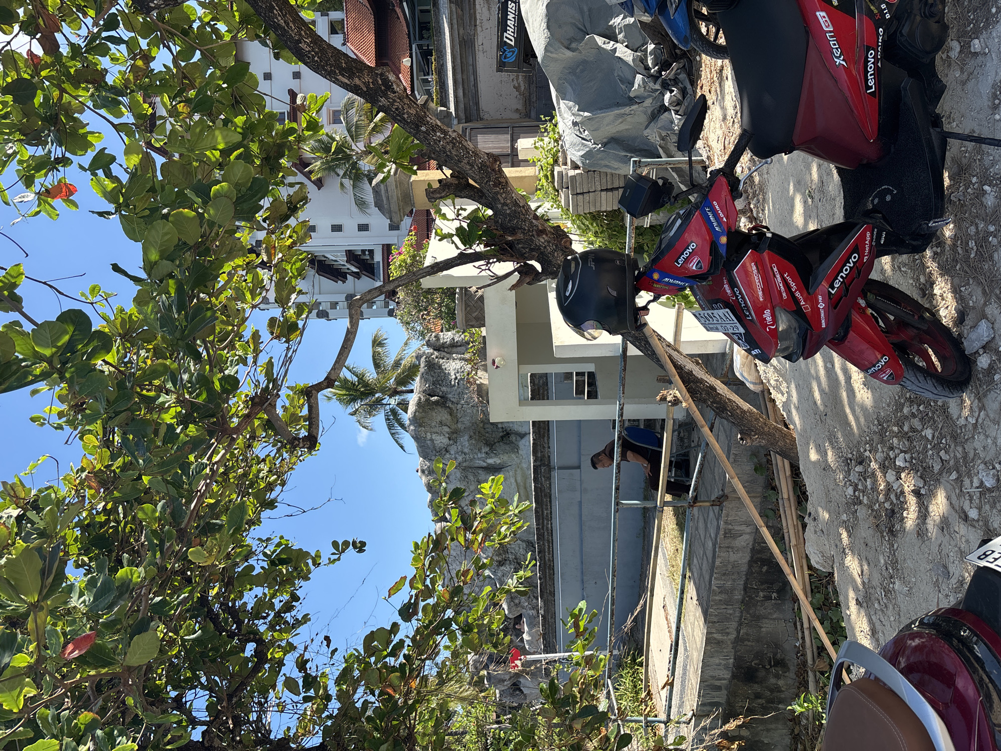 In Kuta Selatan, Indonesia, a red scooter with multiple stickers, including "Lenovo," "Ducati," and "Audi Sport," is parked on a sandy area. A black helmet rests on the handlebars. In the background, a person sits on a chair near a concrete structure and a rock formation. Palm trees and leafy branches overhang the scene, with a white building visible behind the trees and a blue sky above. A maroon scooter is partially visible in the lower left corner.