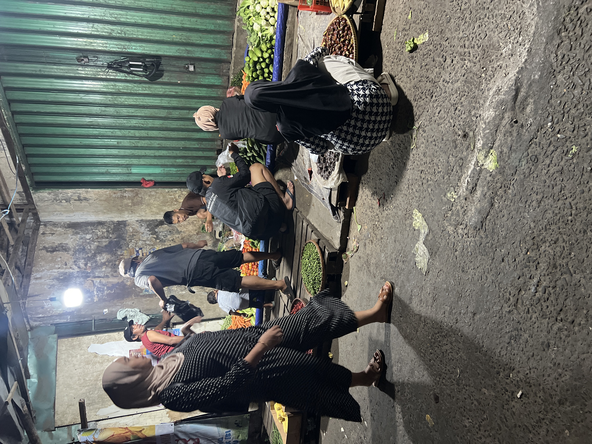 The image depicts a street market scene at night in Citeureup, Indonesia. A woman wearing a black and white striped dress and a light brown hijab walks from left to right across the foreground. Behind her, several people are engaged in activities related to the market. Two men are interacting near a stall with vegetables, one handing a black bag to the other. Another man is seated, looking down. Two women are kneeling near baskets of produce, one wearing a black hijab and patterned clothing, the other a light pink hijab. The environment includes a rough asphalt ground, wooden crates and stalls, and a green corrugated metal shutter in the background. Hanging light bulbs illuminate the scene. Various vegetables such as cucumbers, tomatoes, and leafy greens are displayed.