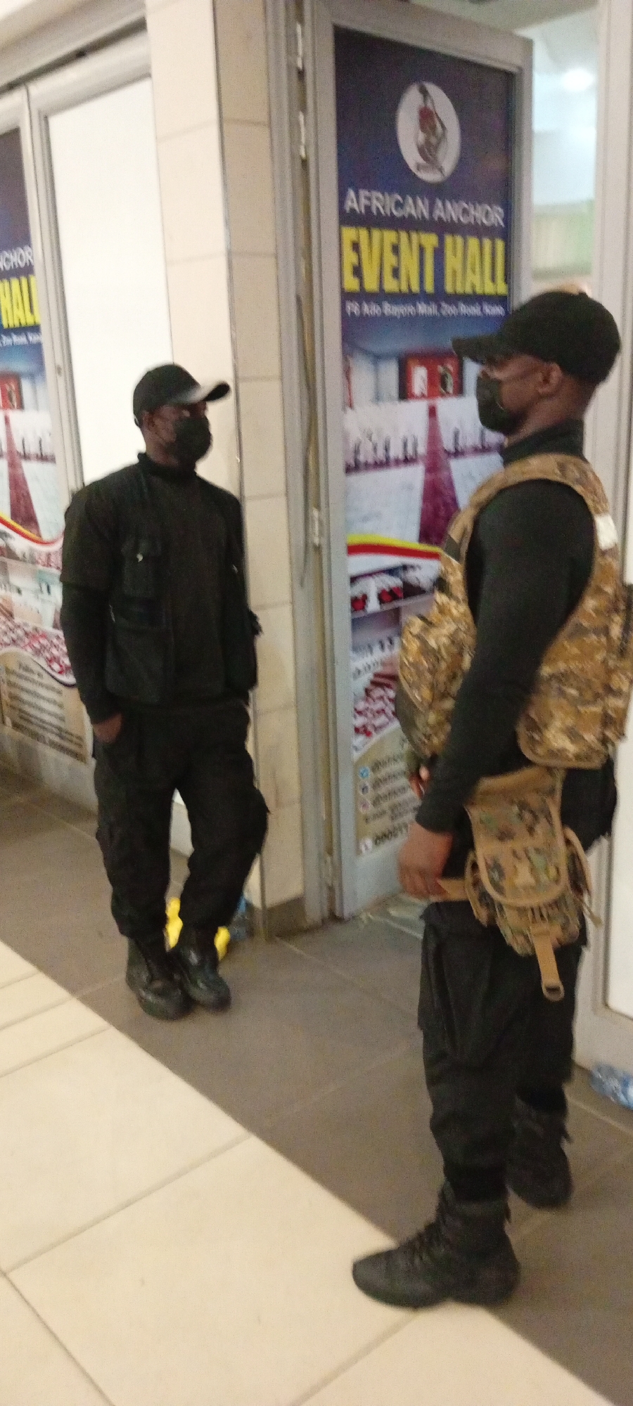 Two men, likely security personnel, stand in front of an establishment. The man on the left wears a black cap, black long-sleeved shirt, and black cargo pants with tactical vest over the shirt, and black boots. He faces towards the right, with his left hand in his pocket. The man on the right wears a black cap, black long-sleeved shirt, black cargo pants, and a camouflage tactical vest with a pouch attached to his belt. He faces towards the left, looking at the other man. Behind them is a glass door with a sign that reads "AFRICAN ANCHOR EVENT HALL" in large yellow letters, with the address "F6 Ado Bayero Mall, Zoo Road, Kano". The environment appears to be an indoor walkway or mall corridor, with tiled flooring. The lighting suggests it is daytime.