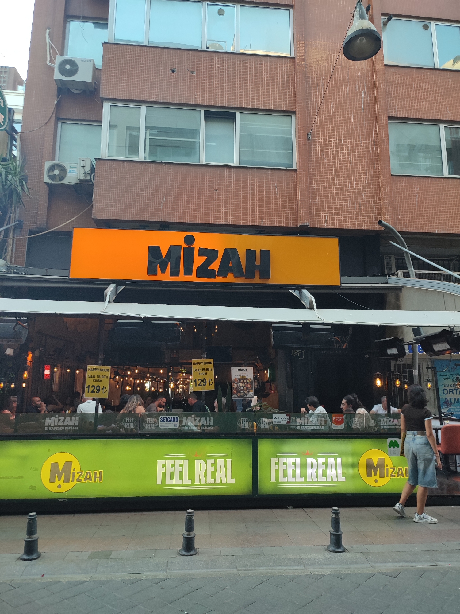 A street-level shot captures the facade of a restaurant named "Mizah" in Kadıköy, Turkey. The restaurant features a prominent orange sign with the name "Mizah" in large black letters. Below the sign, a glass-enclosed area reveals patrons seated inside. Yellow signs advertising "HAPPY HOUR" with a price of "129 ₺" are visible through the glass. The lower section of the building has a bright green illuminated sign with the Mizah logo and the slogan "FEEL REAL." A woman in a denim skirt and a dark top is walking past the front of the restaurant, with her back to the camera. The street in front is paved with gray cobblestones, and two black bollards are positioned on the sidewalk. The building itself is clad in brown brickwork, with several windows on the upper floors. Air conditioning units are mounted on the exterior walls. The overall impression is a lively urban dining establishment.