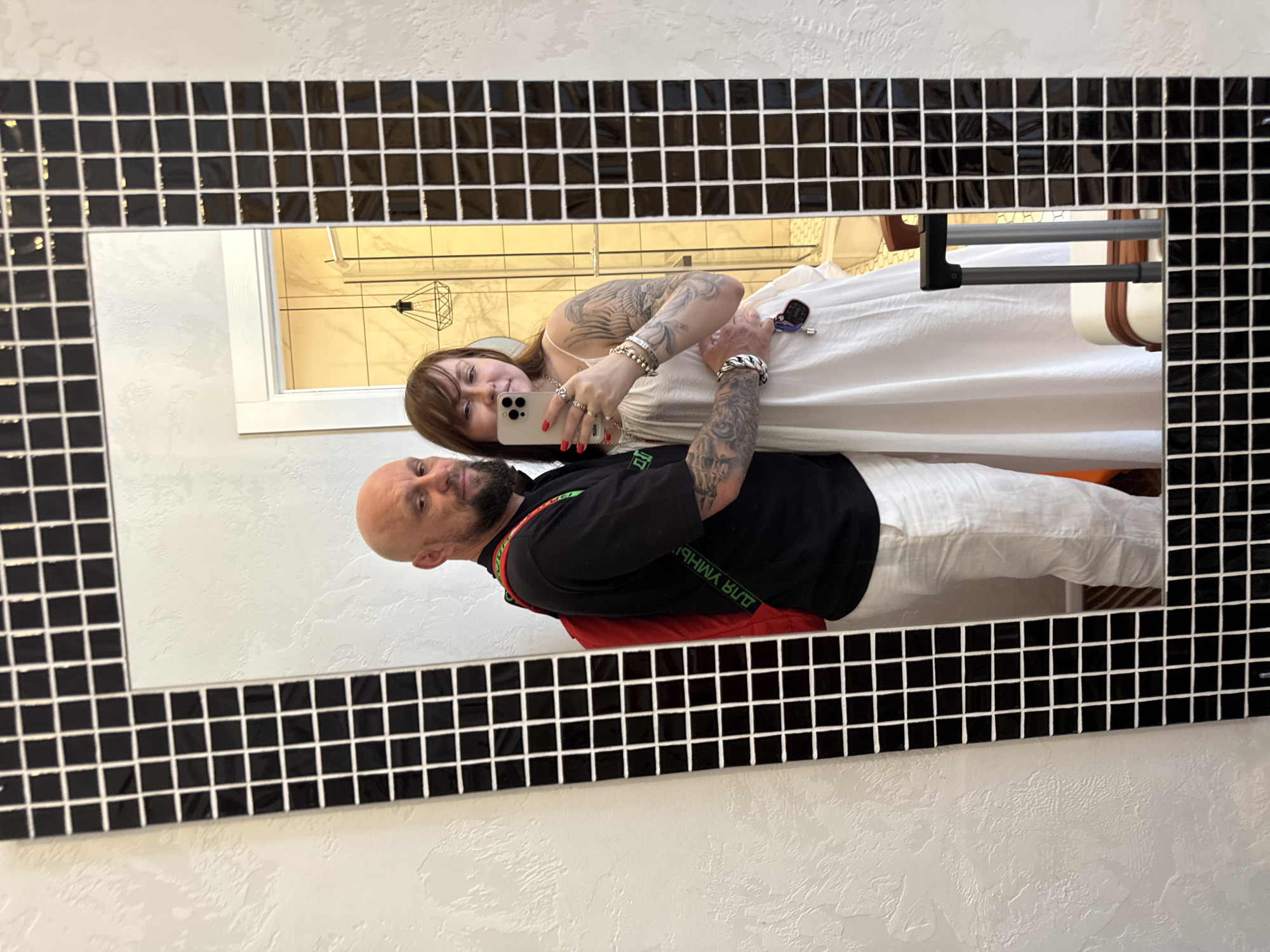 The image is a mirror selfie taken in a bathroom, likely in a hotel, given the presence of a suitcase. A man and a woman are posing for the photo. The man, bald with a beard, is wearing a black t-shirt, white pants, and a red backpack with a green strap. The woman is wearing a white dress and has tattoos on her arms. They are standing close together, with the woman holding a phone to take the picture. In the reflection, a shower stall and a sink are visible, along with a small decorative lamp. The bathroom features a black and white tiled mirror frame and textured white walls. Based on the visual cues, this scene is most likely located in Krasnodar, Russia, a city known for its modern hotel infrastructure and urban lifestyle.