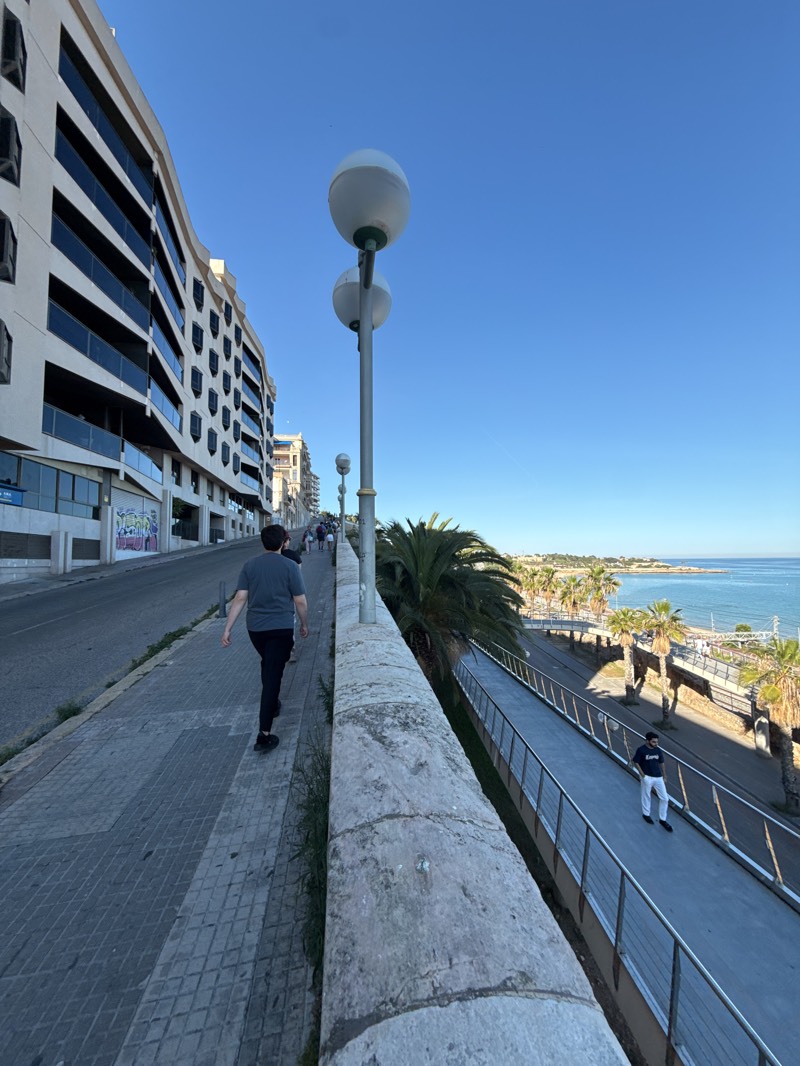 The image shows a scene in Tarragona, Spain. The perspective is from an elevated walkway bordered by a low stone wall. A man in a gray shirt and dark pants walks along the walkway, away from the viewer. To the right of the walkway is a lower pedestrian path with a metal railing, where another man wearing white pants and a dark shirt is standing. Beyond this path are palm trees and the sea.
To the left of the elevated walkway is a multi-story building with dark window panes and what appears to be graffiti on the lower level. Street lamps with two spherical lights each are placed along the walkway. The sky is clear and blue. Other people are walking in the distance.