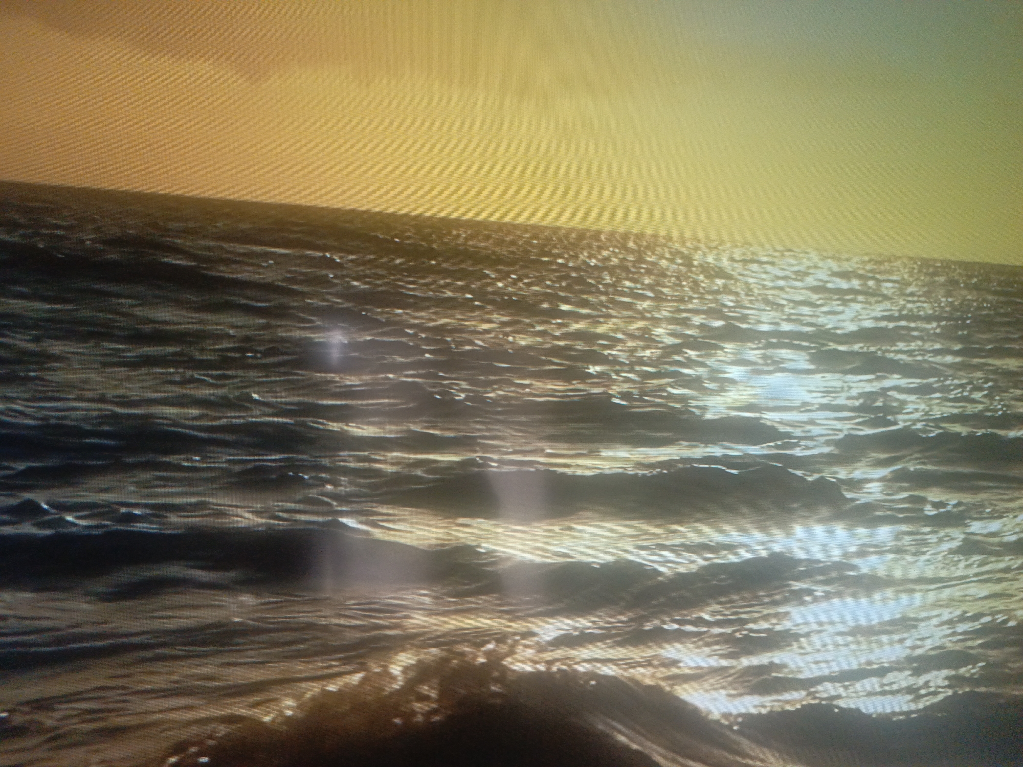 The image depicts an ocean scene, likely during sunset or sunrise, with prominent golden light reflecting on the water's surface. Small waves ripple across the water, creating streaks of light. The horizon line is visible, with a yellowish sky above. The location, Juan Griego, Venezuela, is not discernible from the image itself.