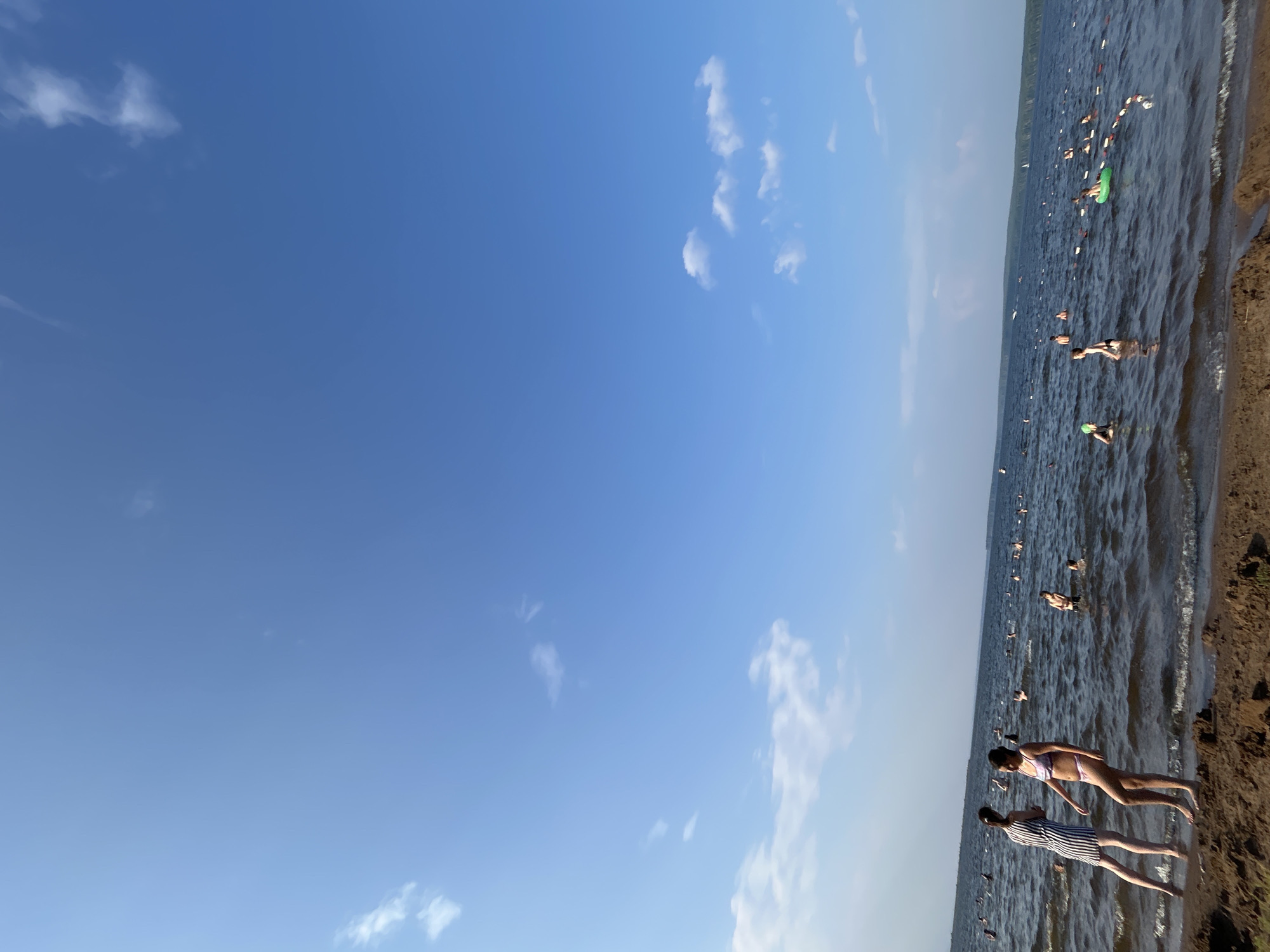The image depicts a wide, calm body of water with many people swimming and wading. Two women are standing on a sandy shore in the foreground, one wearing a striped dress and the other a bikini. The water is dotted with buoys marking a swimming area. In the background, the distant shoreline is lined with trees. The sky is bright blue with scattered white clouds. This scene is consistent with the appearance of the Kama River beach in Chaikovsky, Russia, a popular recreational area during warm weather.