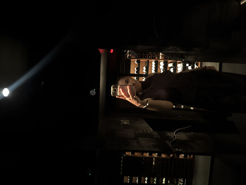 A person takes a selfie in a dark room with a wine rack visible in the background. The room is dimly lit, with a spotlight shining on the left side of the image. The subject is holding a phone in their hand, with a reflection visible in the dark surface below. The overall ambiance is mysterious and dimly lit, drawing attention to the person and the wine rack behind them.