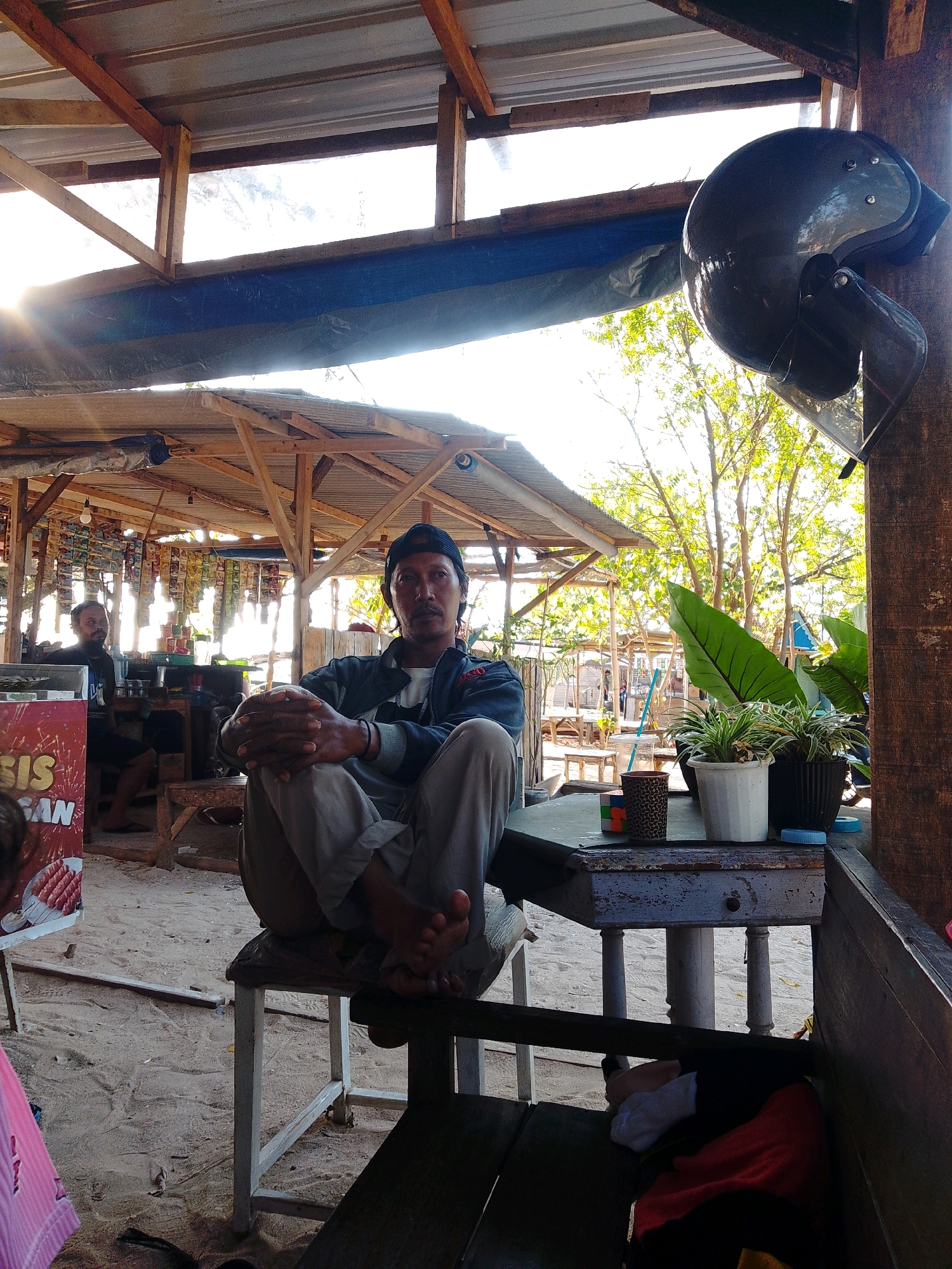 The image depicts an outdoor establishment, possibly a cafe or roadside stall, in Cikal, Indonesia. The man in the foreground is seated on a low wooden bench, facing the camera. He is wearing a blue baseball cap, a dark jacket over a white shirt, and light-colored pants. His hands are clasped in his lap, and his bare feet are visible. Behind him, another man is seated at a table, looking towards the left of the frame. The establishment has a roof supported by wooden beams, with various items, likely food and drinks, hanging from racks in the background. A motorcycle helmet is mounted on a wooden post to the right. Several potted plants are visible on a weathered table in the foreground. The ground is sandy, and trees with lush green foliage are visible in the background, suggesting a tropical environment. Sunlight streams through the roof structure, creating highlights and shadows. The overall impression is of a casual, open-air setting.