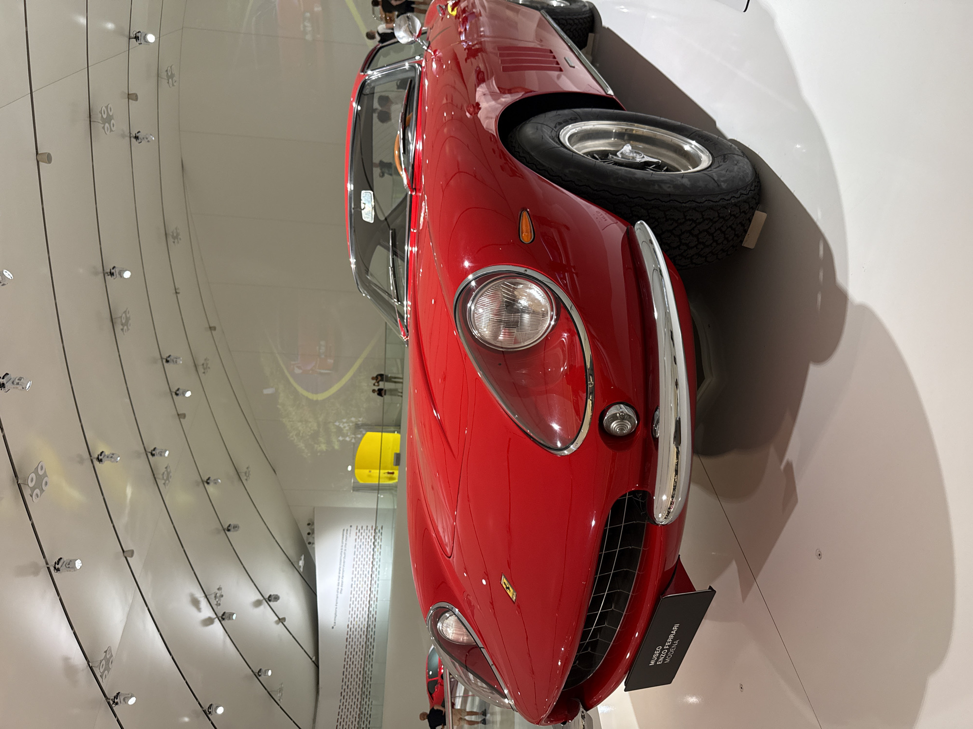 A vibrant red vintage Ferrari is prominently displayed in what appears to be an indoor automotive museum, specifically the Museo Enzo Ferrari in Modena, Italy. The car, a classic two-seater model with elegant curves, chrome trim, and distinct round headlights, is positioned on a light-colored, possibly white or off-white, floor. The image captures the front-right quarter of the vehicle, showcasing its glossy paintwork and a visible tire on the right side.
The setting is characterized by its modern and clean aesthetic. The walls of the exhibition space are strikingly curved and light-toned, adorned with numerous linear elements and small, bright spotlights arranged in rows, creating a sense of dynamic architecture. Reflections of the surrounding environment, including other parts of the exhibition hall, lighting fixtures, and faint figures of people, are visible on the car's polished surface and the glossy floor. In the background, reflections and direct views reveal other museum elements, including a bright yellow object or car and a large white panel with text.
A black display plaque positioned in front of the car clearly states "MUSEO ENZO FERRARI MODENA", confirming the location. Several partial reflections of visitors can be seen on the car's body and in the background, suggesting people are actively viewing the exhibit. The overall ambiance is brightly lit and showcases the vehicle as a prized piece of automotive history.