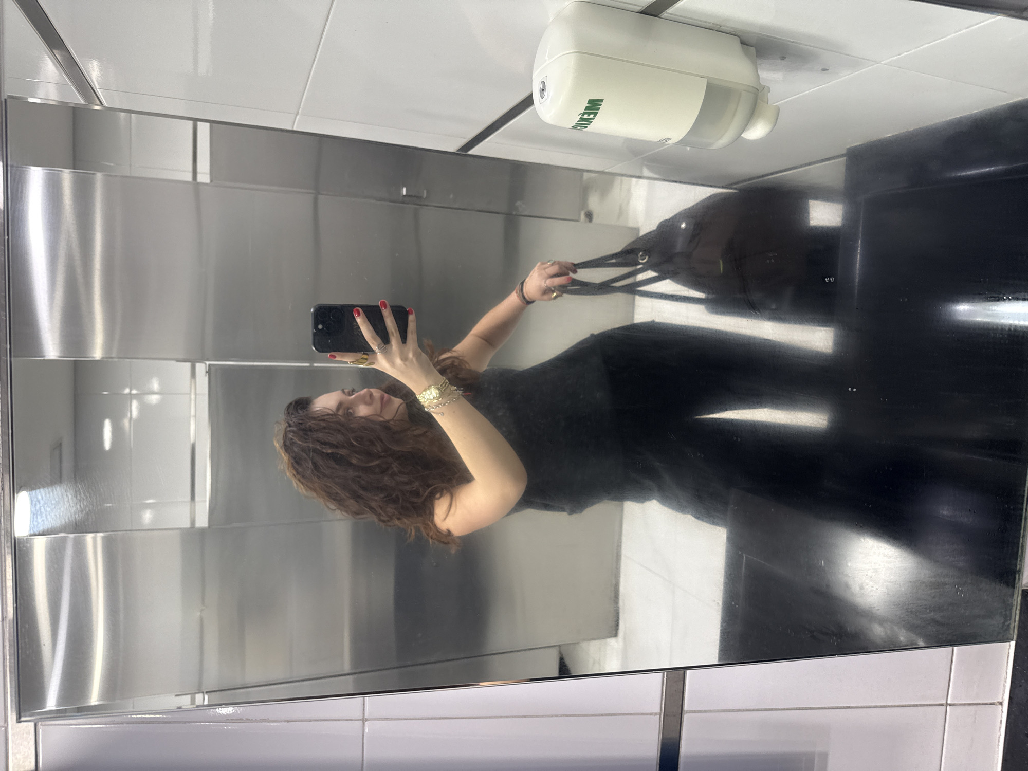 A woman with long, curly brown hair is seen taking a mirror selfie in what appears to be a public restroom or changing area in Mexico City, Mexico. She is reflected from the chest up, wearing a dark sleeveless top or dress. Her right hand, adorned with red nail polish and several rings, holds a black smartphone to capture the image. Her left arm is also visible, featuring a gold-toned watch and bracelets, as she holds the strap of a dark bag, possibly a tote or shoulder bag.
The setting is characterized by its modern and utilitarian design. The background visible in the mirror reflection consists of large, reflective metallic panels, suggesting stainless steel, and white tiled walls. In the upper right portion of the mirror, a white, wall-mounted soap or sanitizer dispenser is visible. The floor, also reflected, appears to be tiled in a light color. The lighting is bright and even, typical of indoor commercial spaces. On the dispenser, the brand name "WEMEX" is clearly legible in green lettering. The woman's expression is not fully discernible, but her pose is typical for taking a mirror selfie.