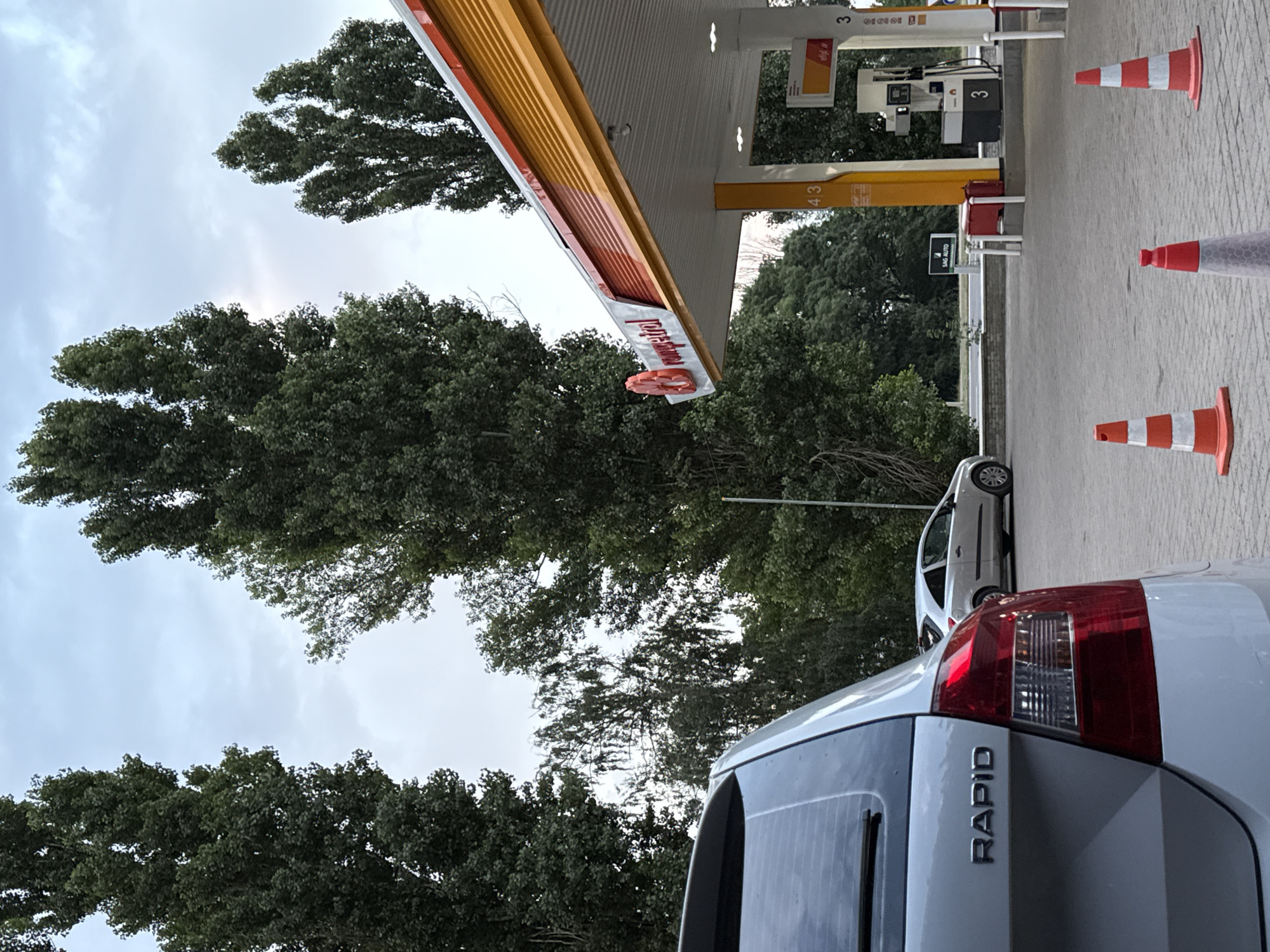 A low-angle shot shows the rear of a white Skoda Rapid car at a gas station in Băcioi, Moldova. The car's rear lights and "RAPID" badge are visible. In the background, a white car is parked next to a gas pump with the number "3" displayed. A yellow and orange gas station canopy, branded "Rompetrol", is visible overhead. Several orange and white traffic cones are placed on the paved ground in front of the gas pumps. Tall, slender trees with dark green foliage line the background, and the sky is overcast.