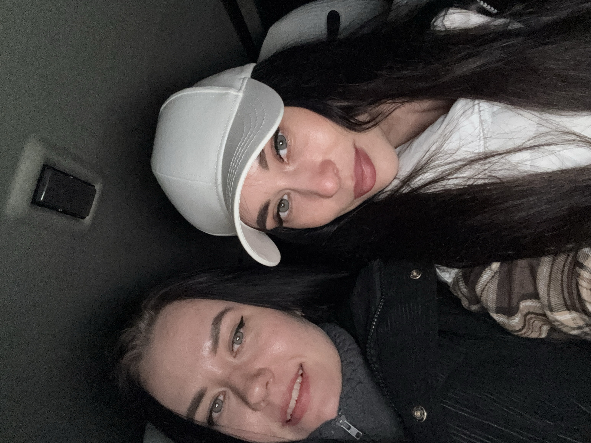 The image shows two women posing for a selfie indoors. The woman at the top is wearing a white cap and a white shirt. She has long, dark hair. The woman at the bottom is wearing a dark jacket over a gray top. The environment appears to be the inside of a vehicle. Based on the available information, the specific location in Chaikovsky, Russia cannot be determined.