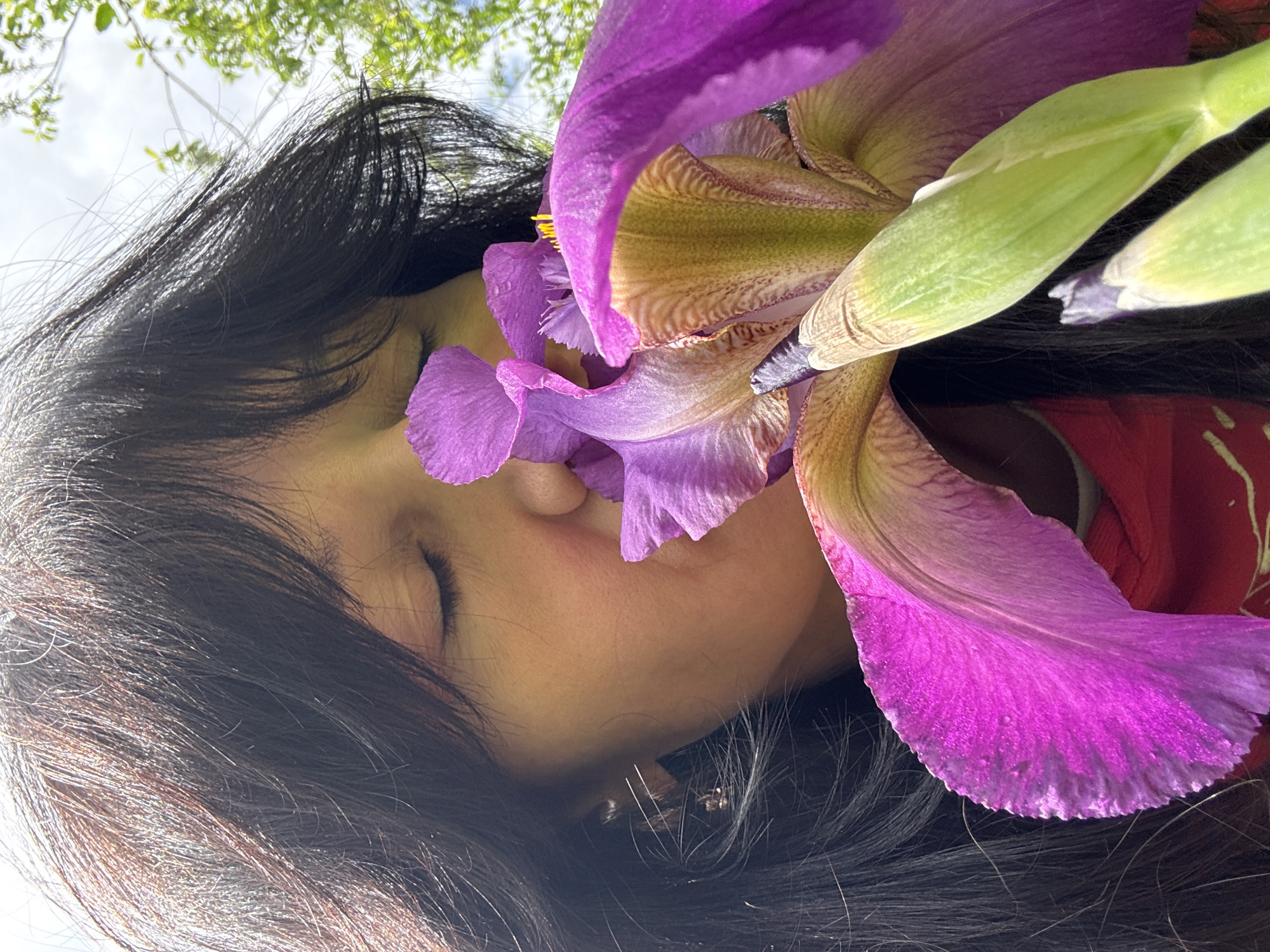 A close-up shot shows a woman's face, partially obscured by a large purple iris. Her eyes are closed, and her dark hair frames her face. A red garment is visible near the edge of the frame. The background includes glimpses of trees and a bright sky. The photograph seems to be taken outdoors during daylight. There is no way to confirm the location of the image being Chaikovsky, Russia.
