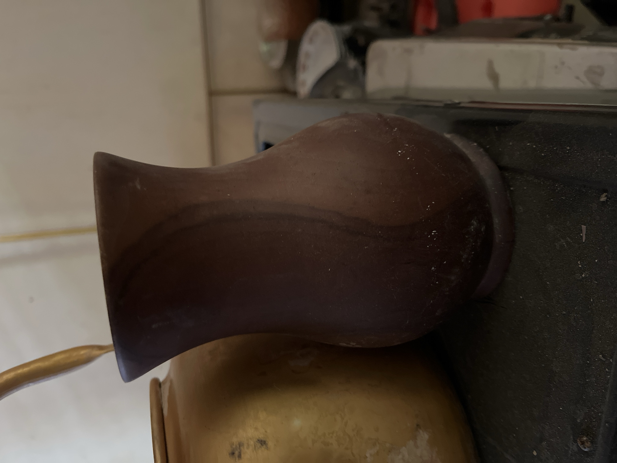 A close-up, eye-level shot shows a dark brown, gourd-shaped vase with a narrow neck and flared opening. The vase is positioned to the right of a larger, gold-colored pot with a rounded body and a narrow rim, partially visible on the left side of the frame. A thin, golden object, possibly a straw or spoon, emerges from the top of the vase. The surfaces of both the vase and the pot appear dusty, with visible specks of white on the dark vase. The background is out of focus, revealing a tiled wall and various household items, suggesting an indoor setting, likely a kitchen or storage area. The scene is illuminated by ambient light. No individuals are present in the image. The exact location in Kota Kuala Simpang, Indonesia is not ascertainable from the visual information provided.