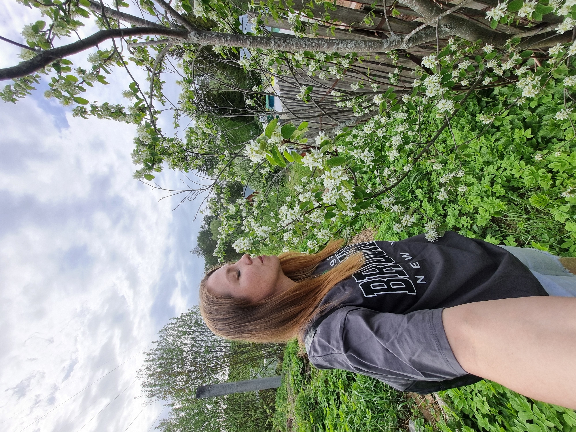 The image depicts a woman lying on her back in a grassy area. She has long brown hair and is wearing a grey t-shirt with the word "BROOKLYN" printed on it. Her eyes are closed. The background includes a tree with white blossoms and a wooden fence. The sky is overcast with clouds. The image's location is Chaikovsky, Russia.