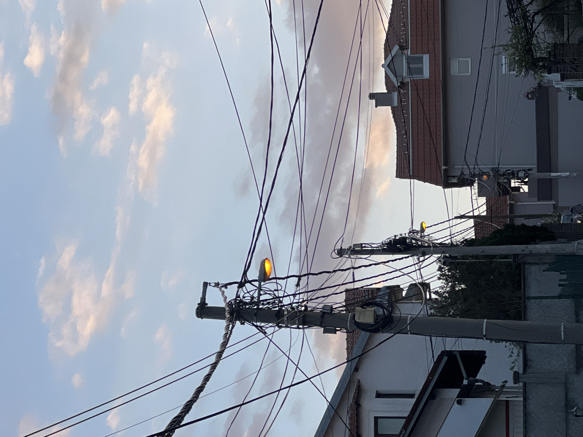 A daytime, low-angle shot shows a typical urban scene in Palilula Urban Municipality, Serbia, cluttered with power lines. The sky is filled with clouds, tinged with pastel hues suggesting either sunrise or sunset. Utility poles, laden with a dense web of black wires and electrical equipment, dominate the foreground. One pole has a lit street lamp. Residential buildings with tiled roofs and light-colored walls can be seen in the background. The overall impression is one of urban density and infrastructure.