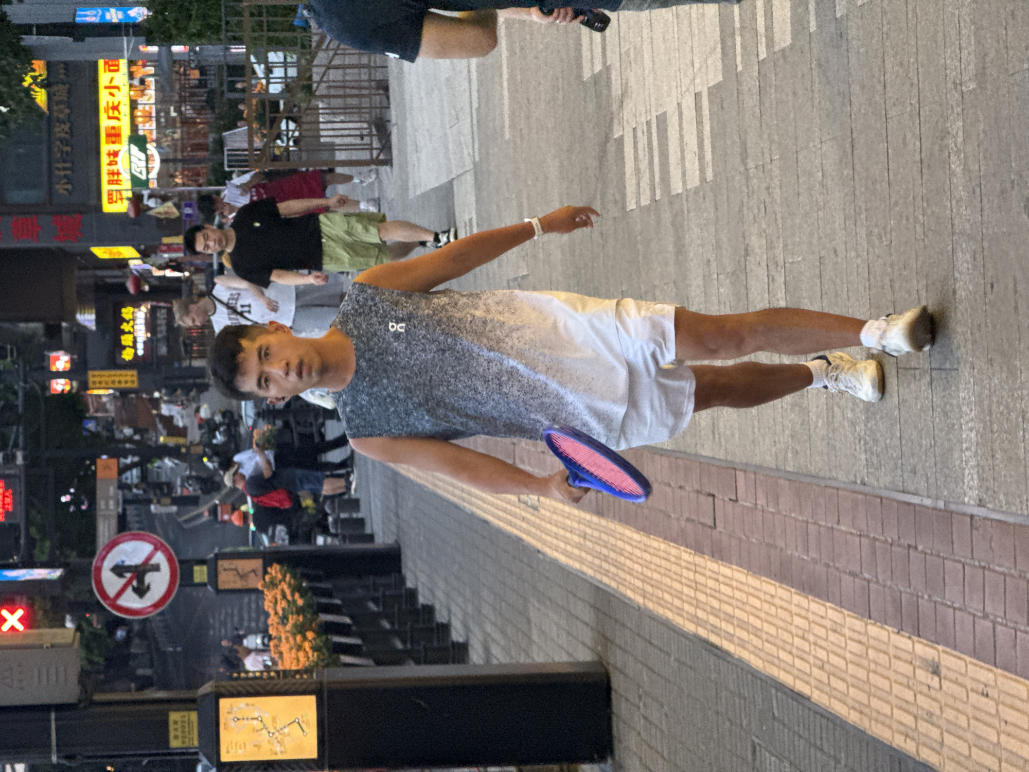 A young East Asian man with short black hair is walking towards the camera on a city sidewalk in Yuzhong District, China. He is wearing a sleeveless grey and white speckled tank top, white shorts, white socks, and white athletic shoes. In his right hand, he holds a bright pink and blue paddle tennis racket. He is wearing a white wristband on his left wrist.
The sidewalk is paved with textured yellow and brown bricks, with a raised tactile paving strip to the left of the man. Behind him, other pedestrians are visible, including a man in a black t-shirt and green shorts and a person in a white tank top and grey shorts. The background features a street with businesses displaying illuminated signs in Chinese characters, and traffic signals. An orange traffic signal with a red 'X' symbol is visible above a street sign. The lighting suggests it is late afternoon or early evening.