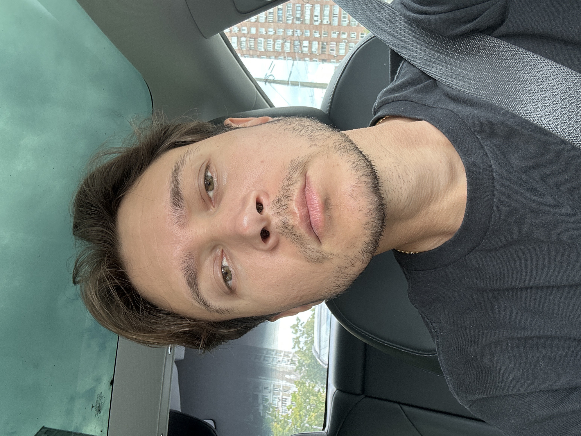 The image shows a selfie taken from inside a car, with the driver wearing a black t-shirt and a gold chain. The driver's face is visible from the chest up, with short brown hair and a light stubble beard. The background includes the car's interior, with black leather seats and a seatbelt. Through the car window, tall brick buildings and trees are visible, characteristic of an urban environment in Kyiv, Ukraine. The sky is overcast.