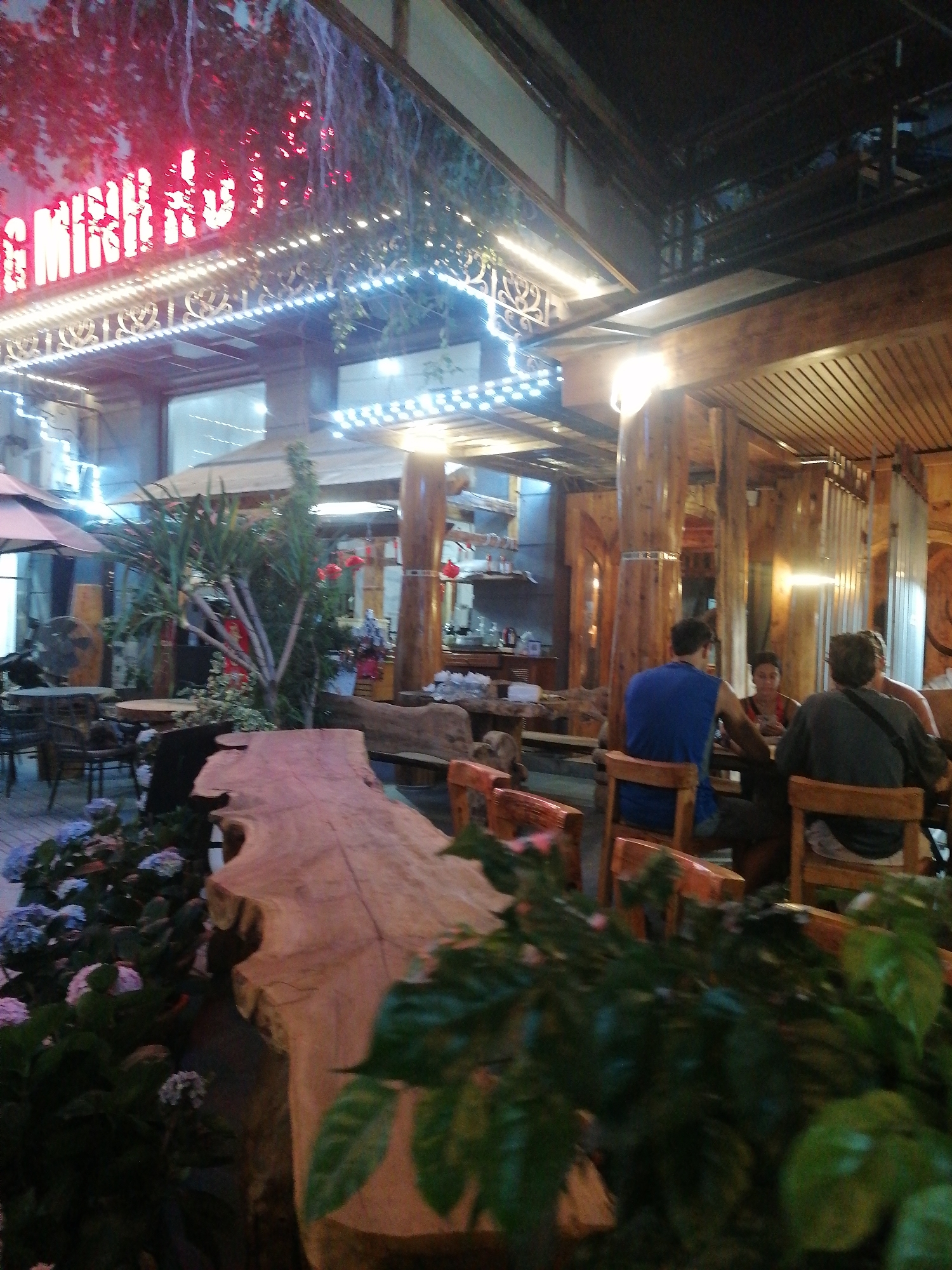 The image depicts an outdoor seating area of a restaurant or establishment in Bai Chay Ward, Vietnam. Overhead, a red neon sign is visible, possibly spelling out "Minh", along with decorative lighting. The building appears to be constructed with wooden beams and pillars, giving it a rustic look. Three people are seated at a table within the seating area. One wears a blue tank top. The area is decorated with plants, including flowering bushes in the foreground, and a large, unfinished wooden slab sits nearby. More tables with chairs and umbrellas are seen on a patio.