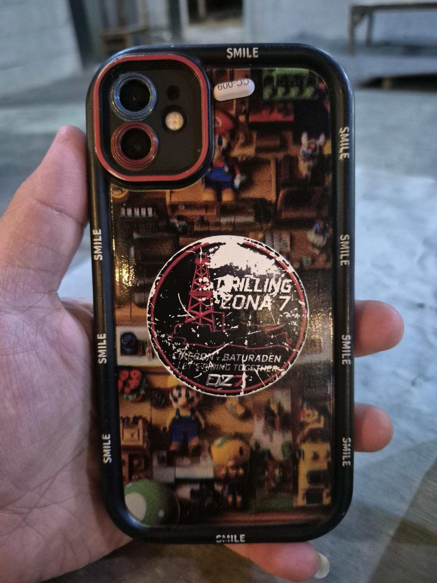 A person's hand holds a black smartphone with a colorful case featuring a collage of images, including cartoon characters, and a circular sticker with text that reads "TRILLING ZONA 7." The sticker is worn and scratched. Along the sides of the phone case, the word "SMILE" is printed repeatedly. A small white sticker with black text "600-009" is visible near the top edge of the phone. The background appears to be an outdoor setting, possibly a street or sidewalk, with a blurred background of muted colors and what looks like a wooden structure in the distance. The lighting suggests it is daytime. The image was taken in Pabelan, Indonesia.